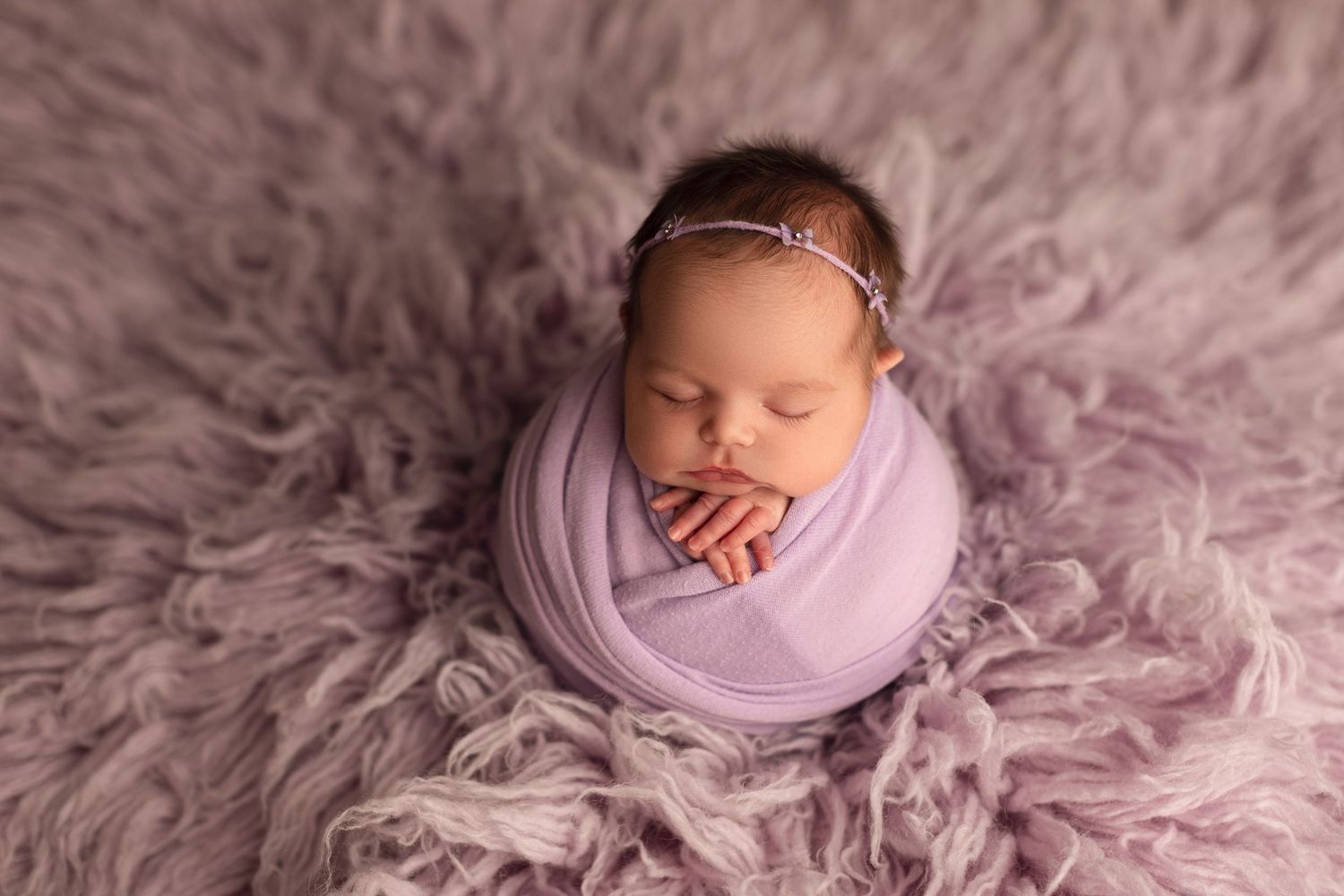 Newborn baby girl wrapped in lavender swaddle sleeping peacefully on a soft purple fluffy rug.