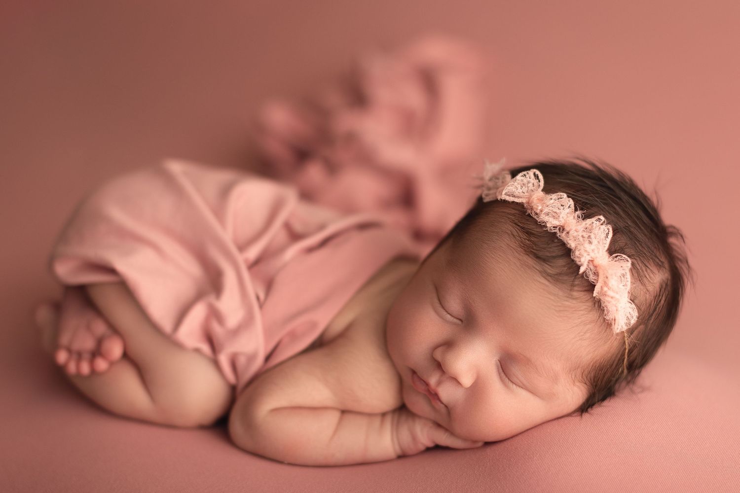Sleeping newborn baby girl in pink wrap and floral headband on a soft pink backdrop.