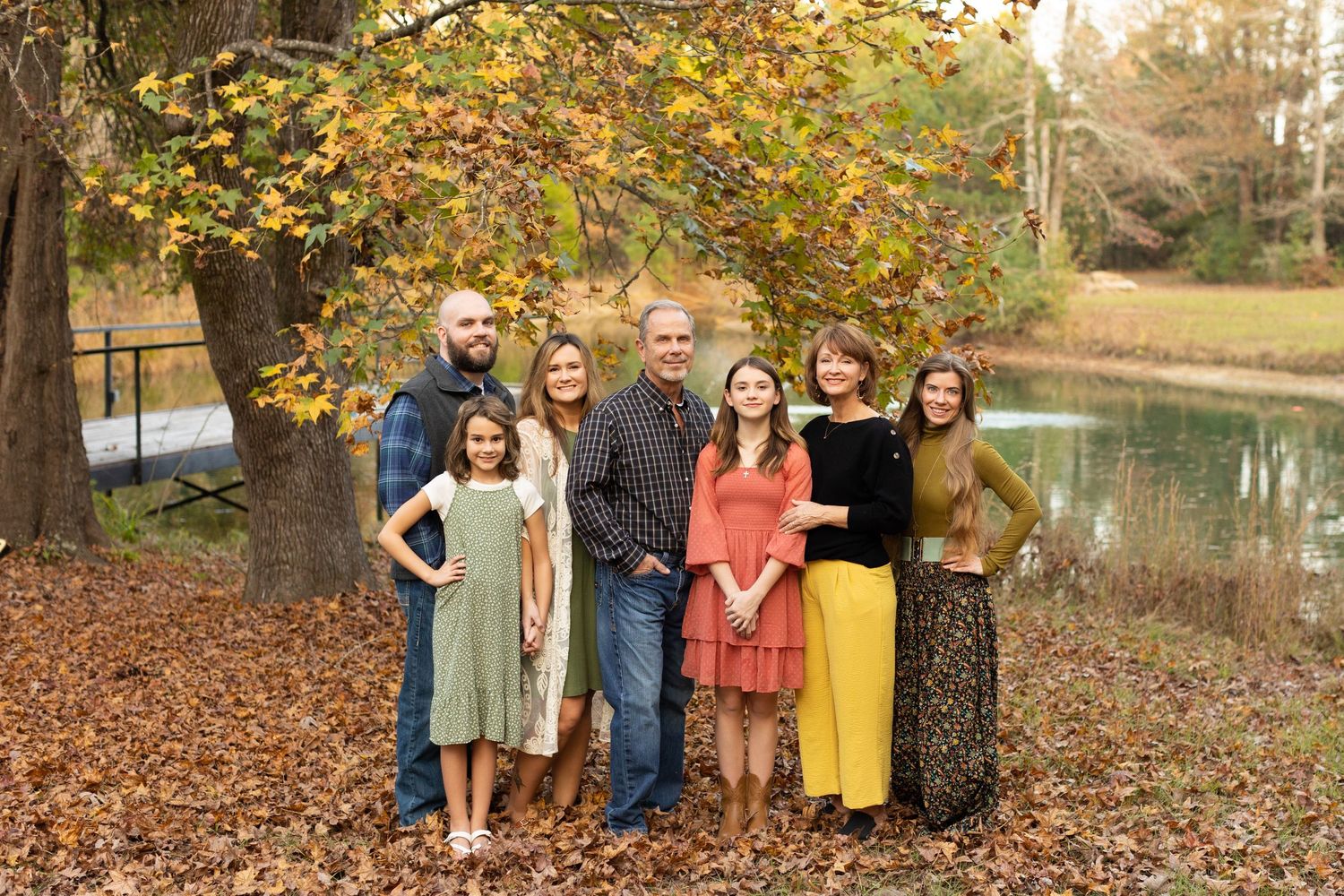 Family of seven posing outdoors under colorful autumn trees near a pond during fall family photo session. Family photographer Atlanta TX