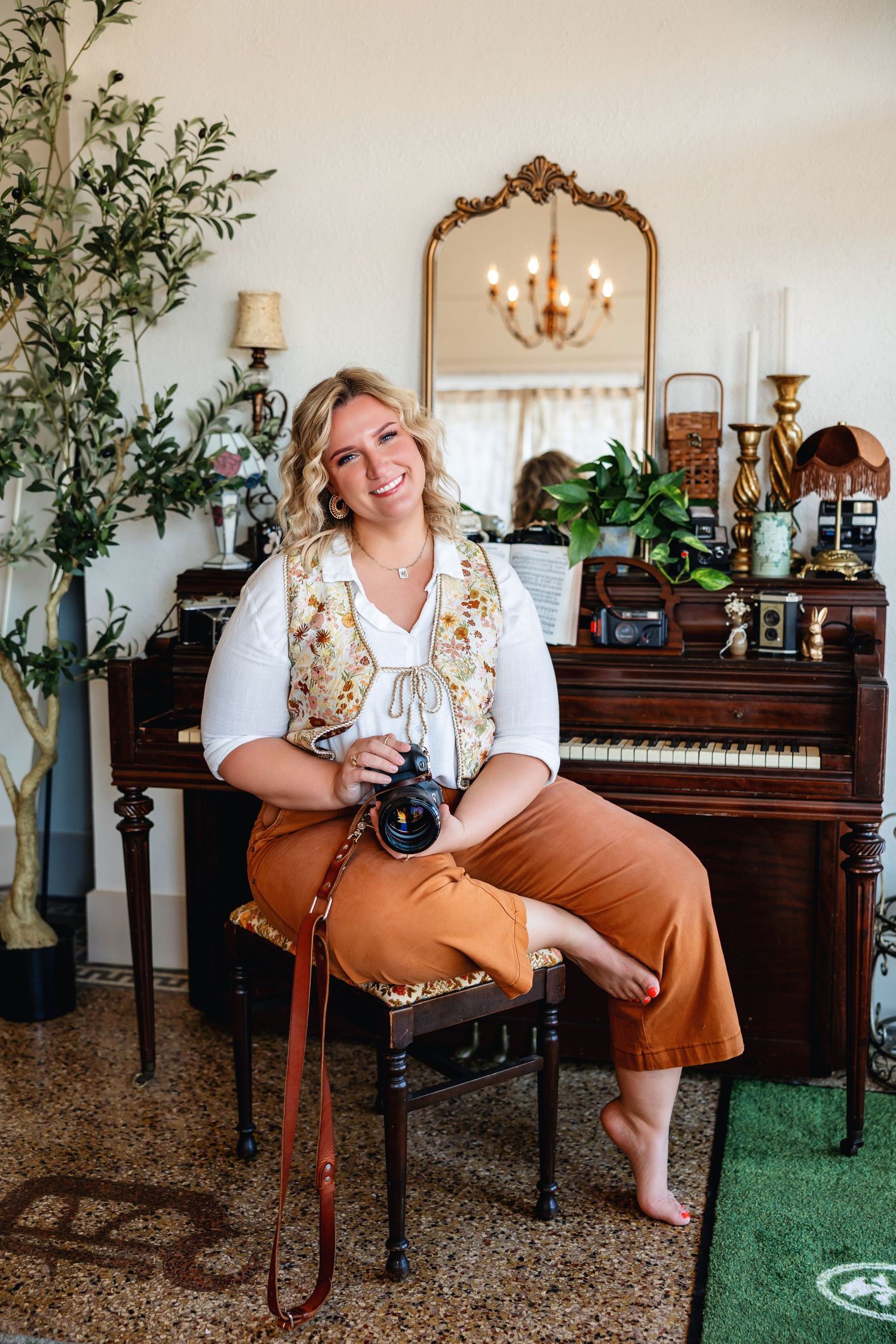 Smiling blonde woman holding a camera sits barefoot near a vintage piano with gold mirror and antique decor.