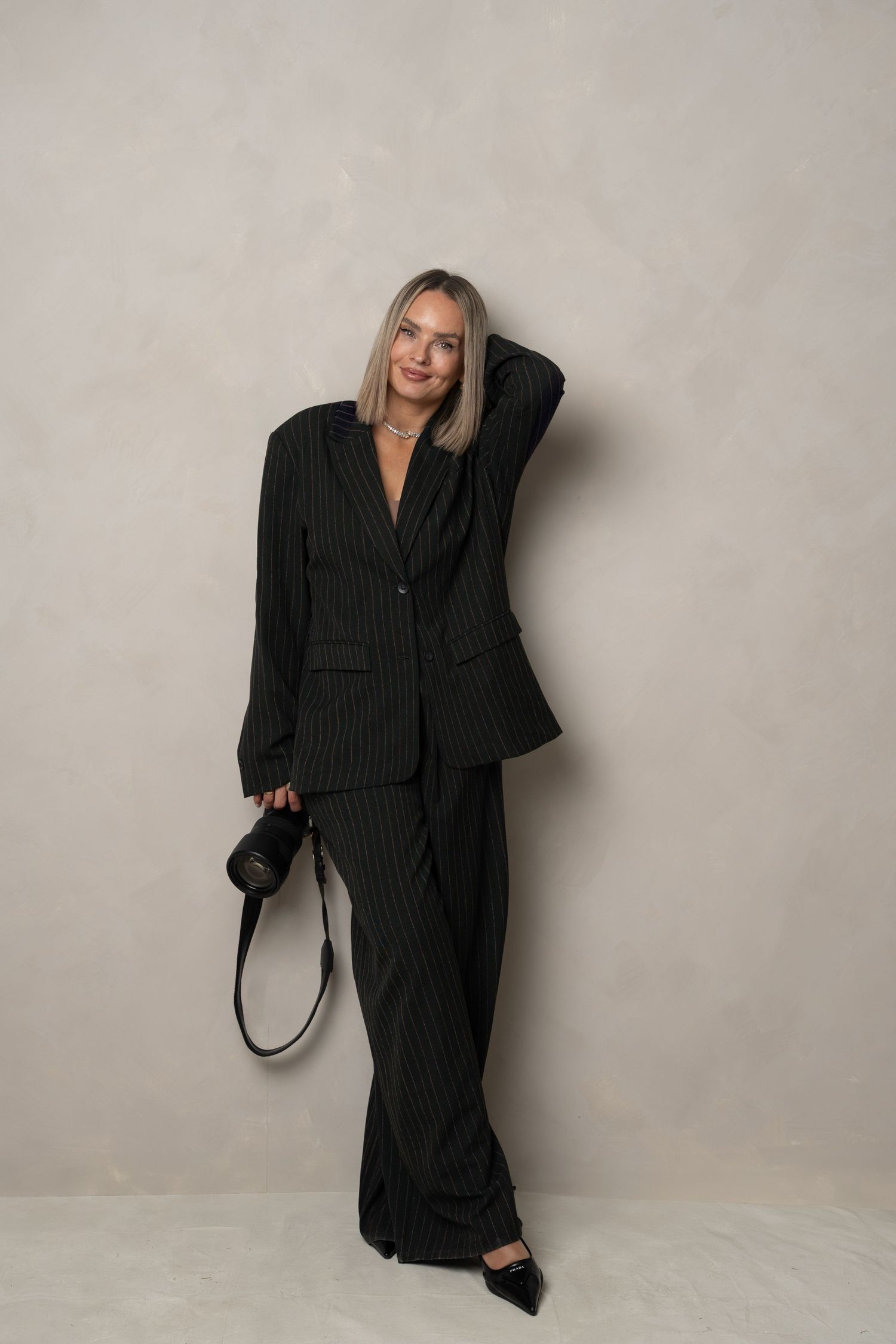 A stylish woman in a black pinstripe suit poses casually against a light colored wall with a camera strap.