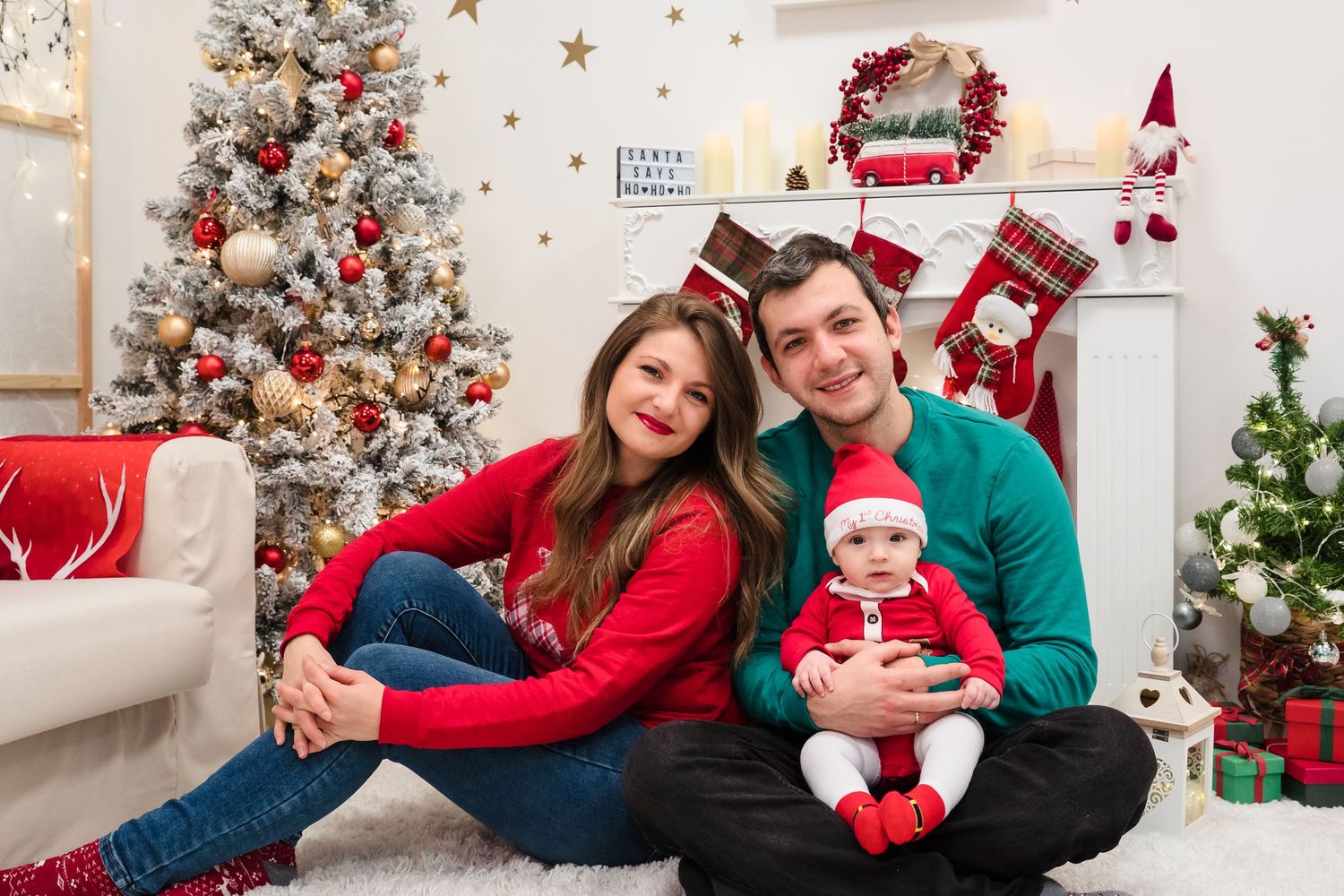 A smiling couple in red and green sweaters holds a baby in a Santa hat on a cozy rug in a festive Christmas photo studio.