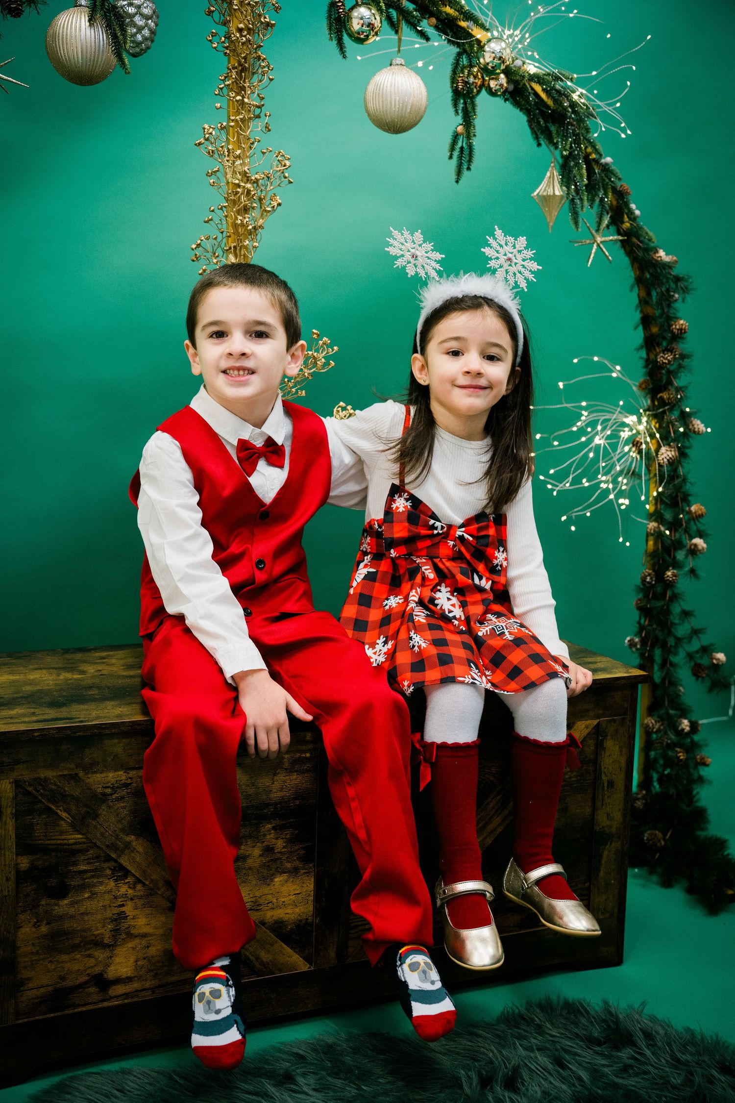 Young boy in a red vest and bow tie sits beside a girl in a plaid dress on a wooden trunk against a green holiday backdrop.