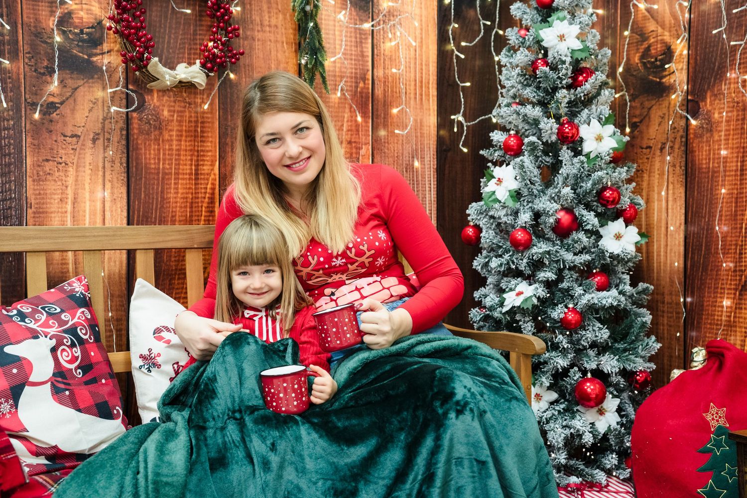 Smiling mother and young daughter in red sweaters sip hot drinks under a green blanket beside a Christmas tree.