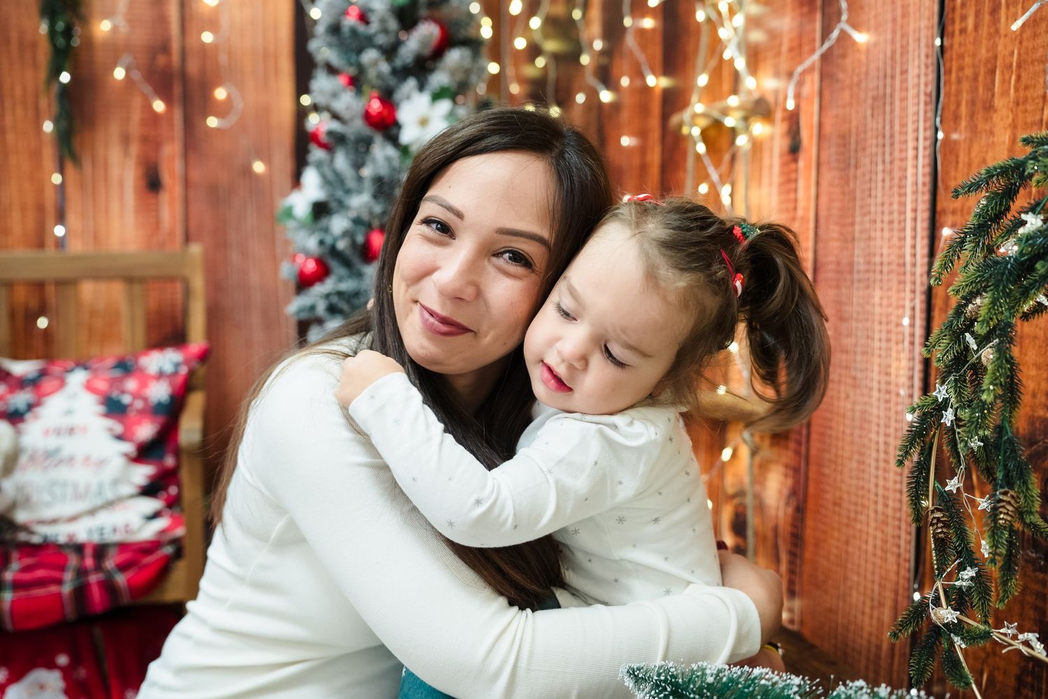 Smiling mother in a white sweater hugs her little daughter in front of a Christmas tree and wooden wall with lights.