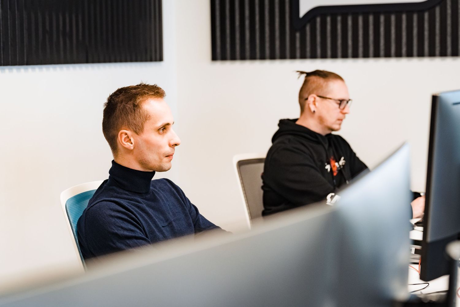 Two support team members sit at office desks, looking at their computer screens in a bright modern workspace.