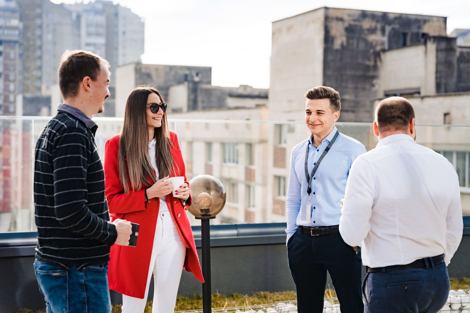 Four coworkers enjoy coffee and conversation on a rooftop terrace with a city view on a sunny day.
