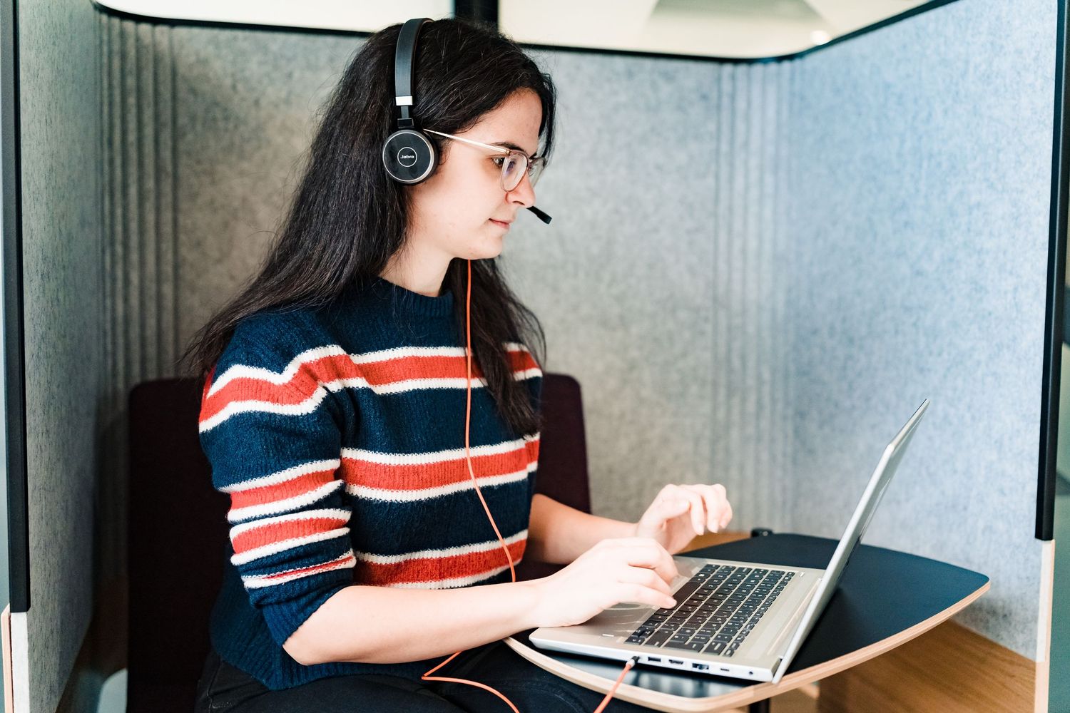 Young woman in a striped sweater and headset types on a laptop inside a soundproof office pod with gray acoustic walls.