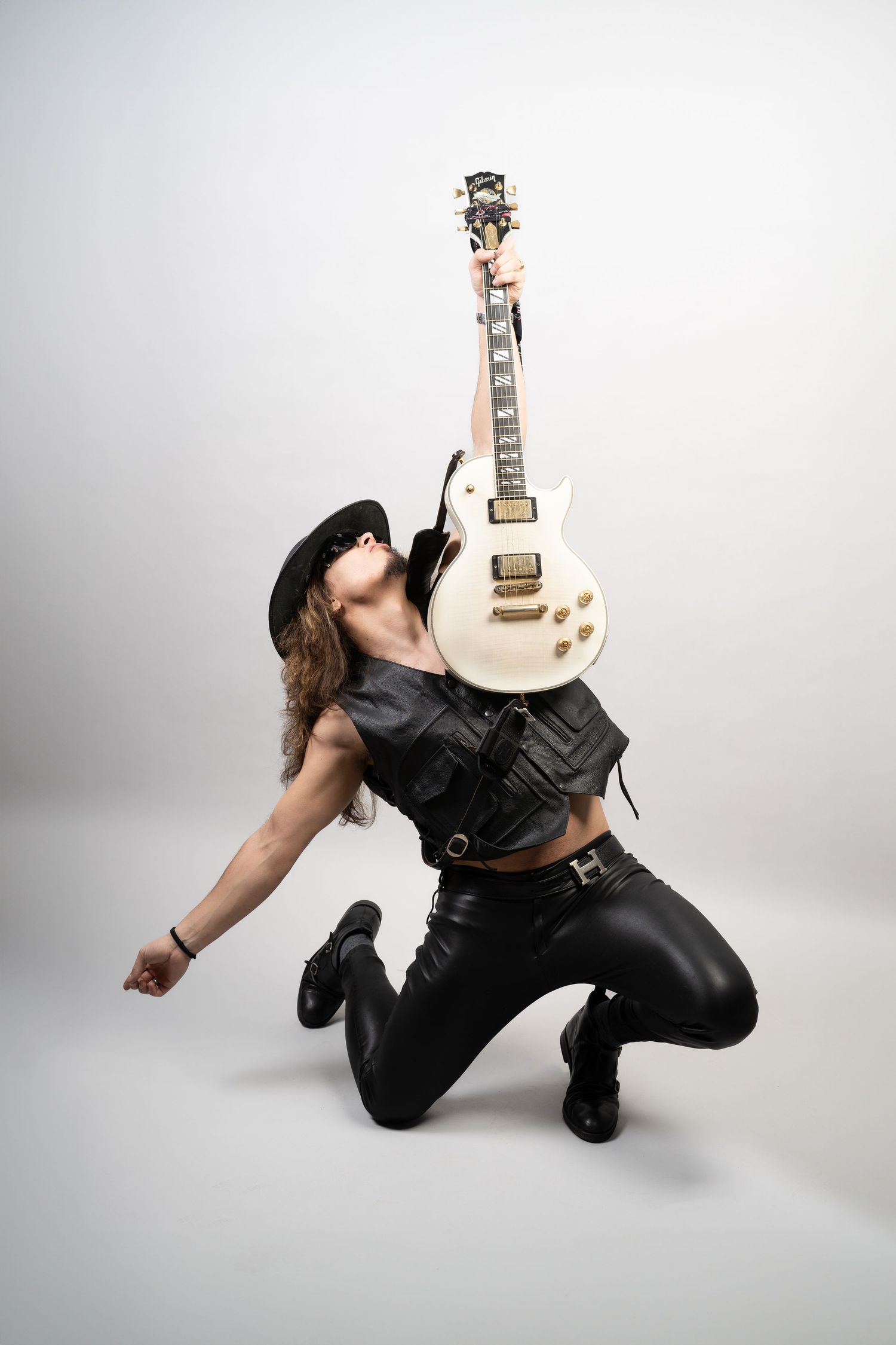 Rock guitarist in a black hat and leather vest kneels and lifts a white electric guitar overhead against a bright studio background.