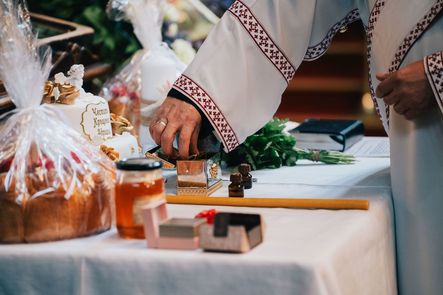 Priest in white vestments with embroidered trim blesses an Easter table with baked bread, honey jar, herbs and small candles.
