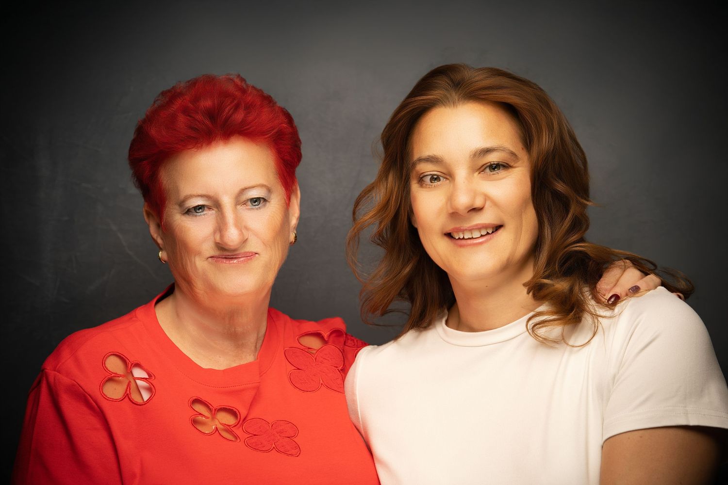 A red-haired woman in a red top and a young woman in white pose together smiling against a dark studio background.