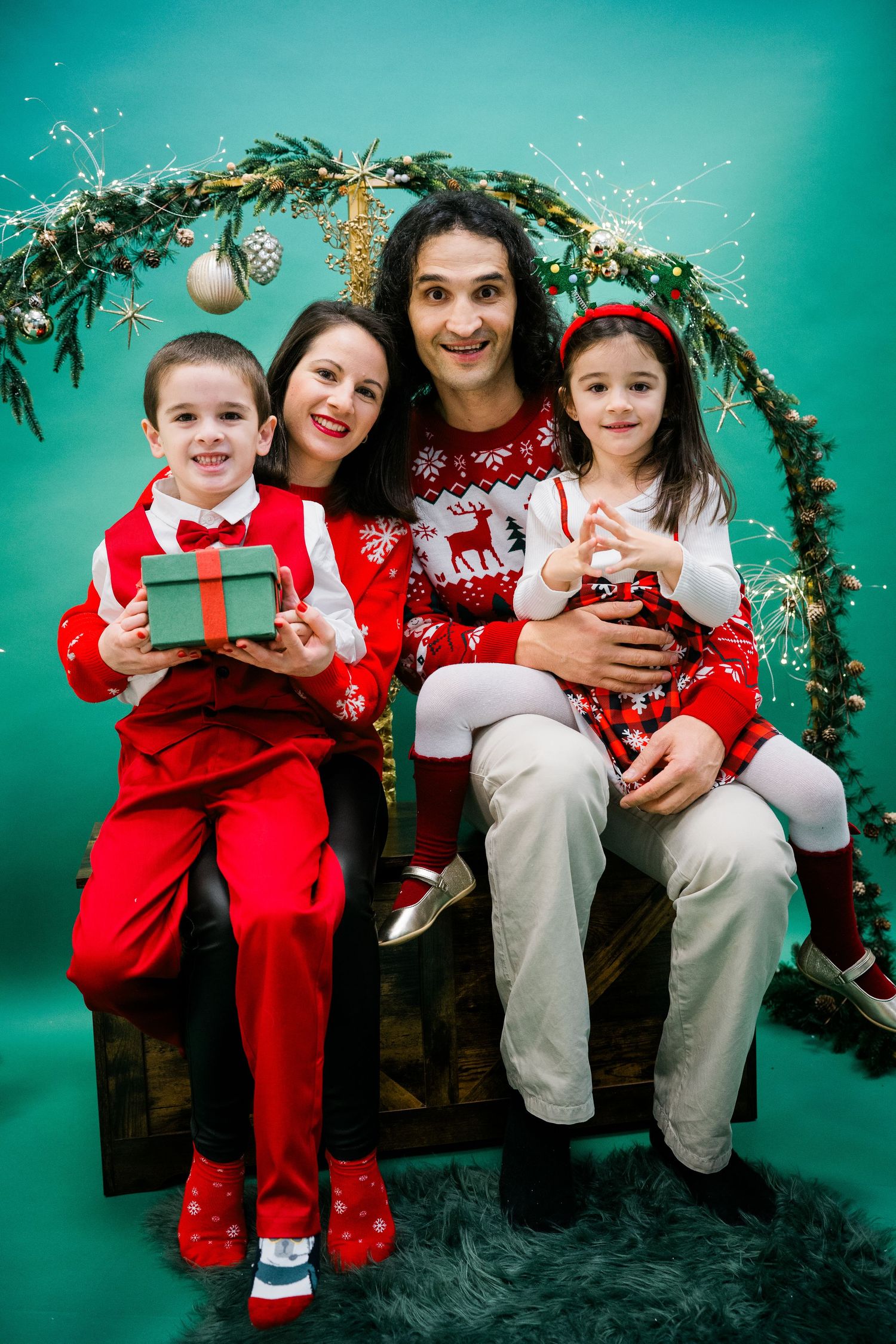 Smiling family of four in red holiday outfits posing together on a wooden bench with a green wreath arch and Christmas gift.