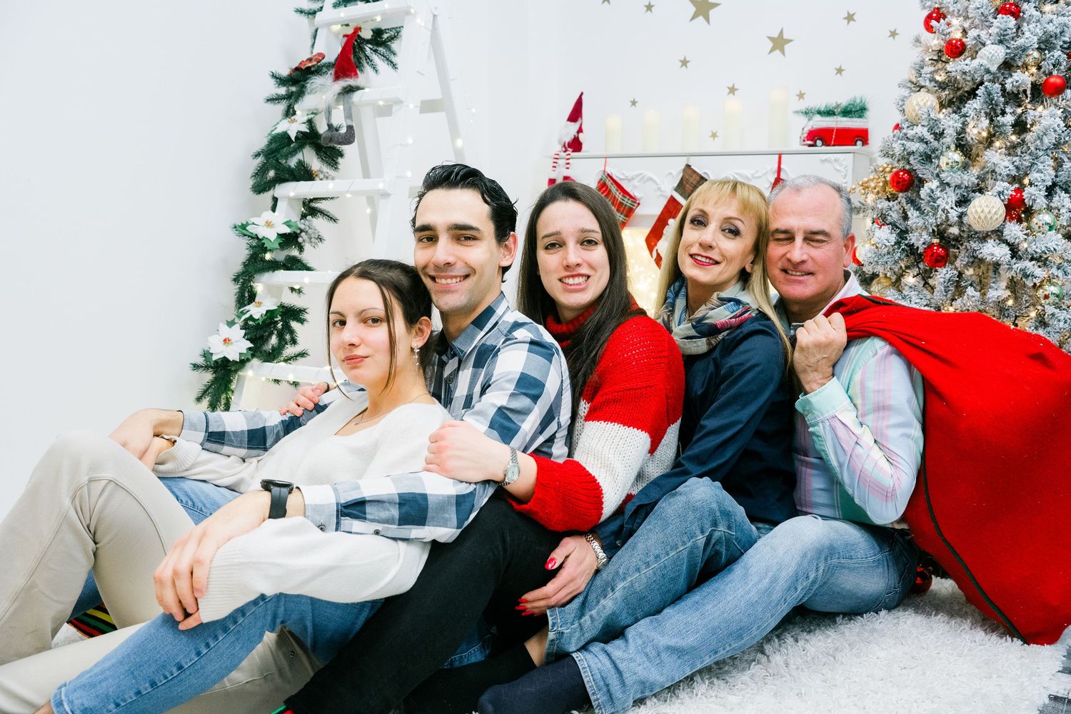Cheerful family of five relaxes on a white rug by a decorated Christmas tree with a red Santa gift sack.