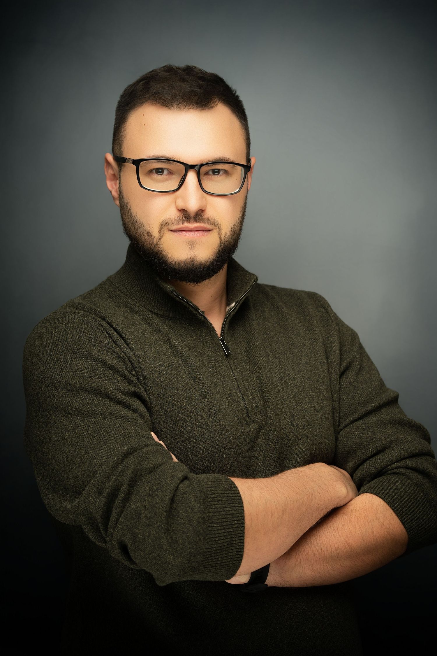 Professional headshot of a bearded man in glasses and an olive green zip sweater posing with arms crossed against a dark gray background.