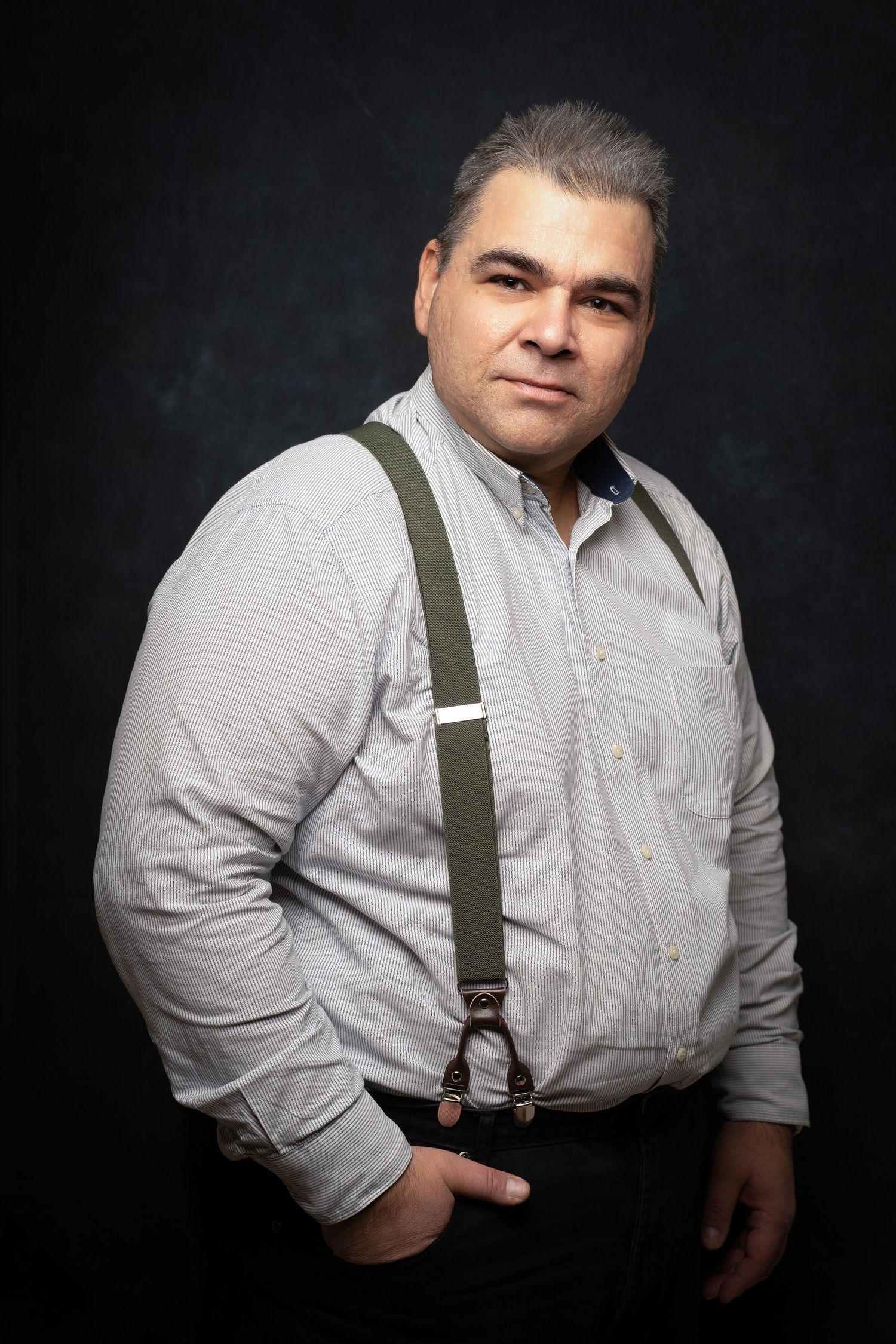 Man with gray hair in a light checkered shirt and olive suspenders posing with a hand in his pocket against a dark textured backdrop.