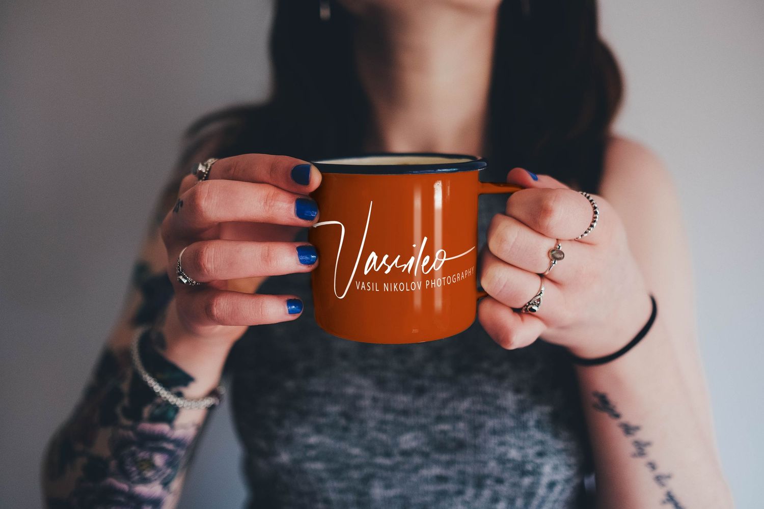 A woman with tattooed arms and stacked silver rings holds up an orange enamel mug with white branding against a gray background.