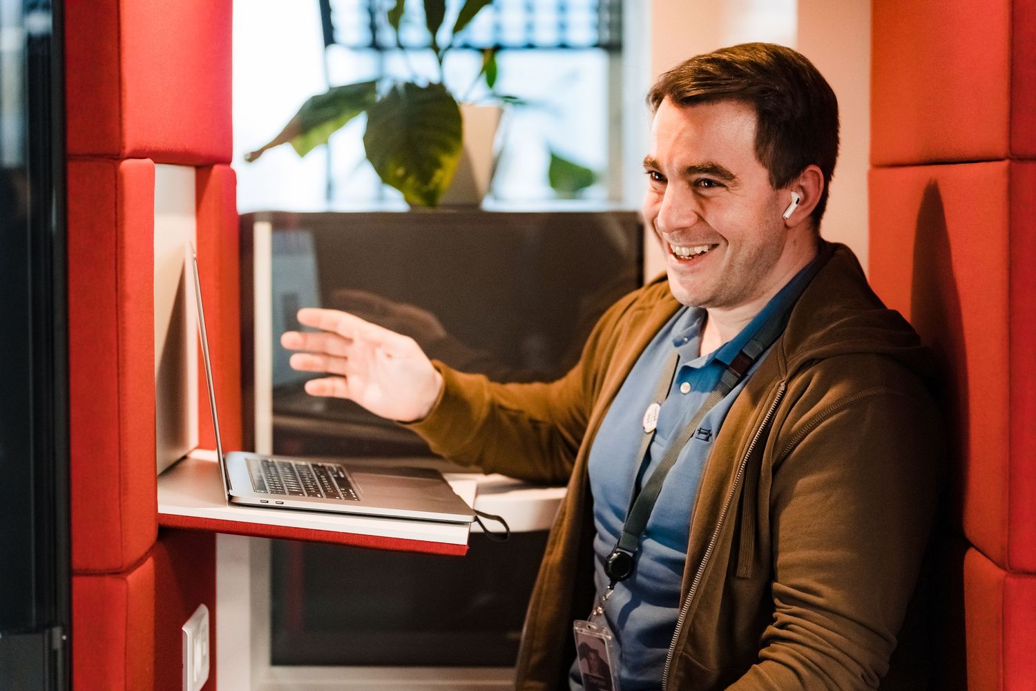 Cheerful man in a brown cardigan gestures while chatting during a video call at a laptop inside a red office booth.