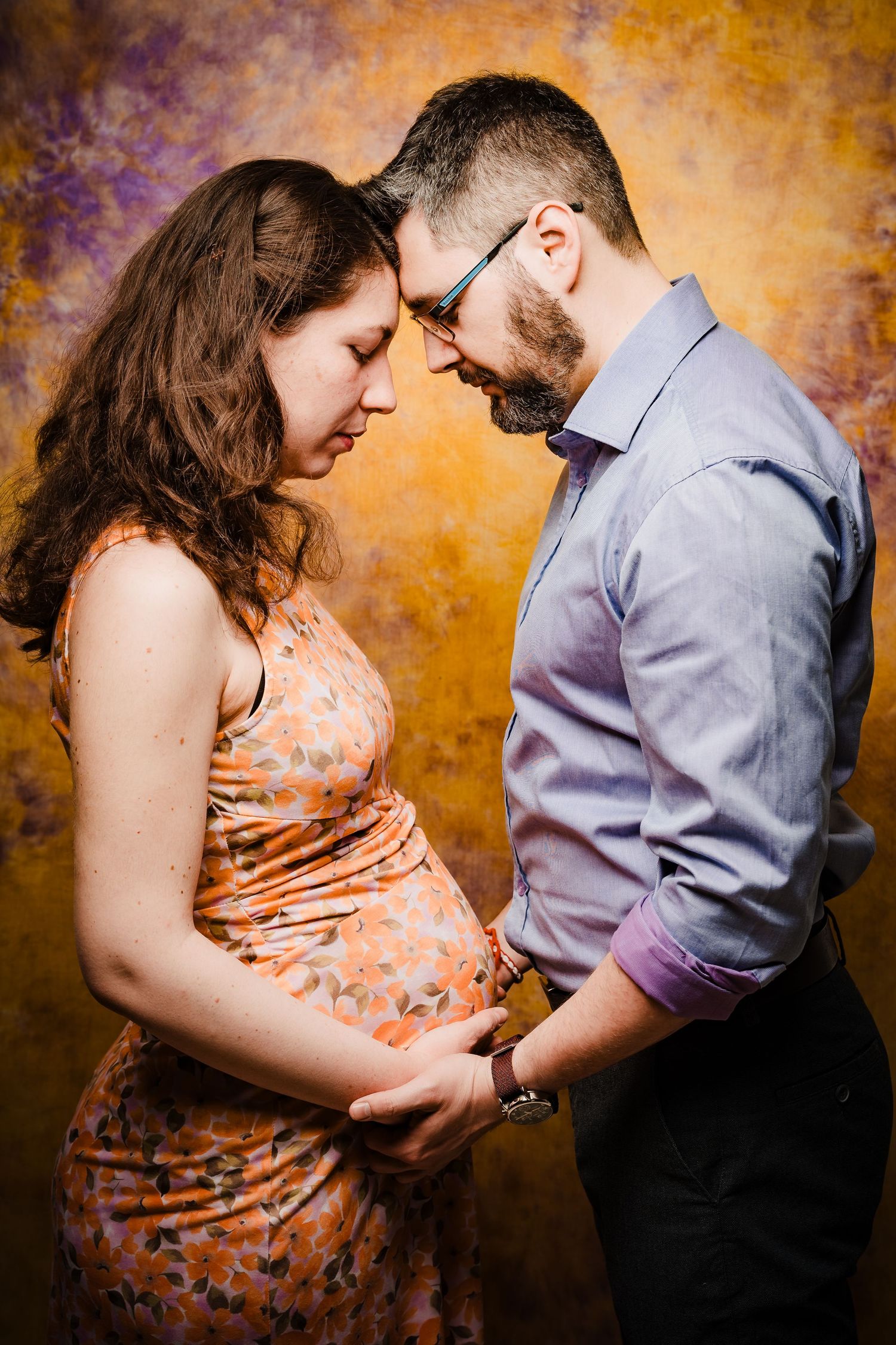 Expectant couple touch foreheads in profile, hands resting on the baby bump, against a colorful abstract golden backdrop.