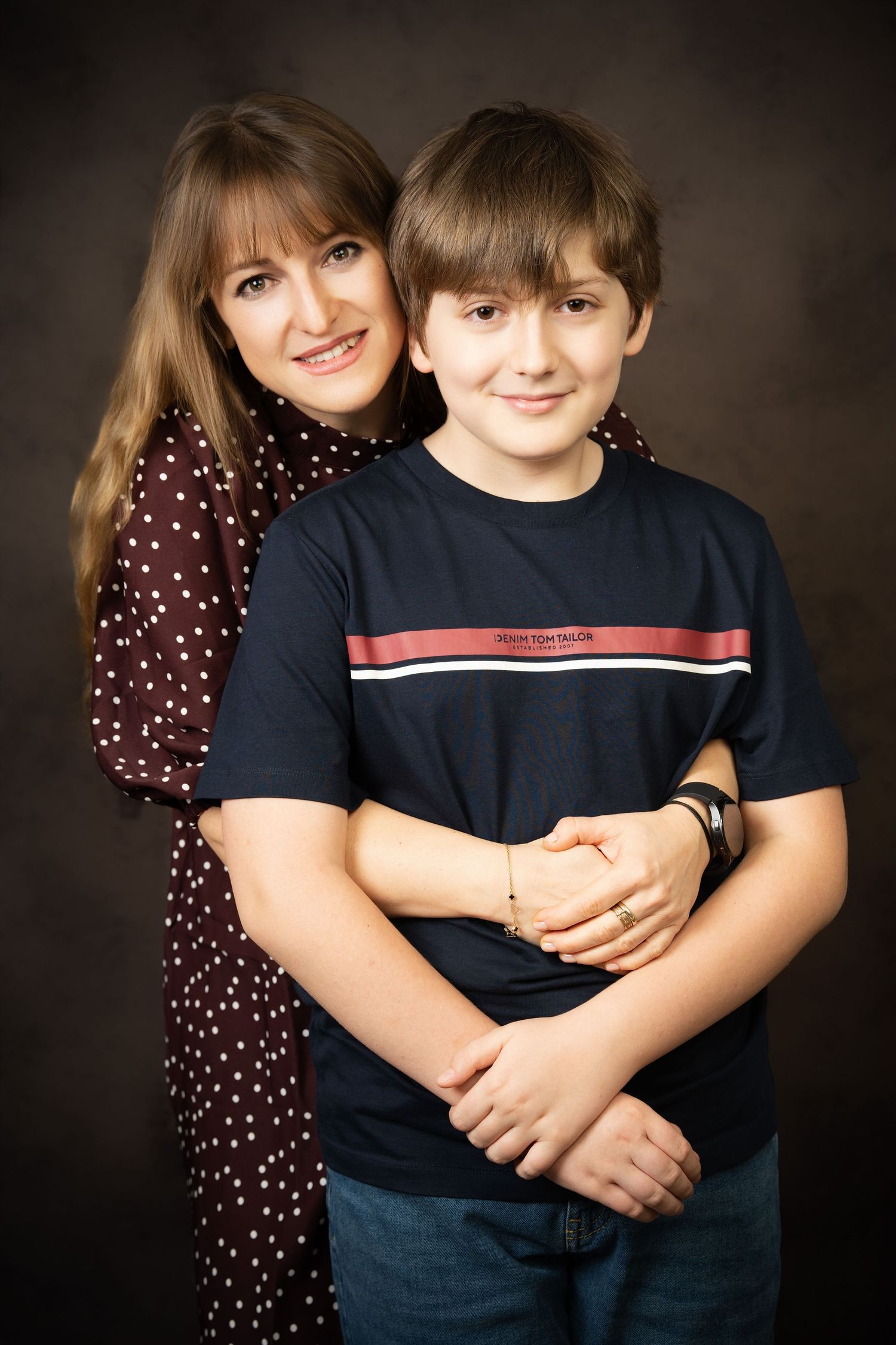 A smiling woman in a polka dot blouse hugs a boy in a navy striped t-shirt from behind in a studio portrait.