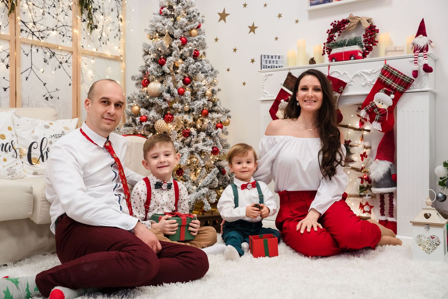A family of four in matching red and white outfits poses on a fluffy rug beside a decorated Christmas tree and fireplace.
