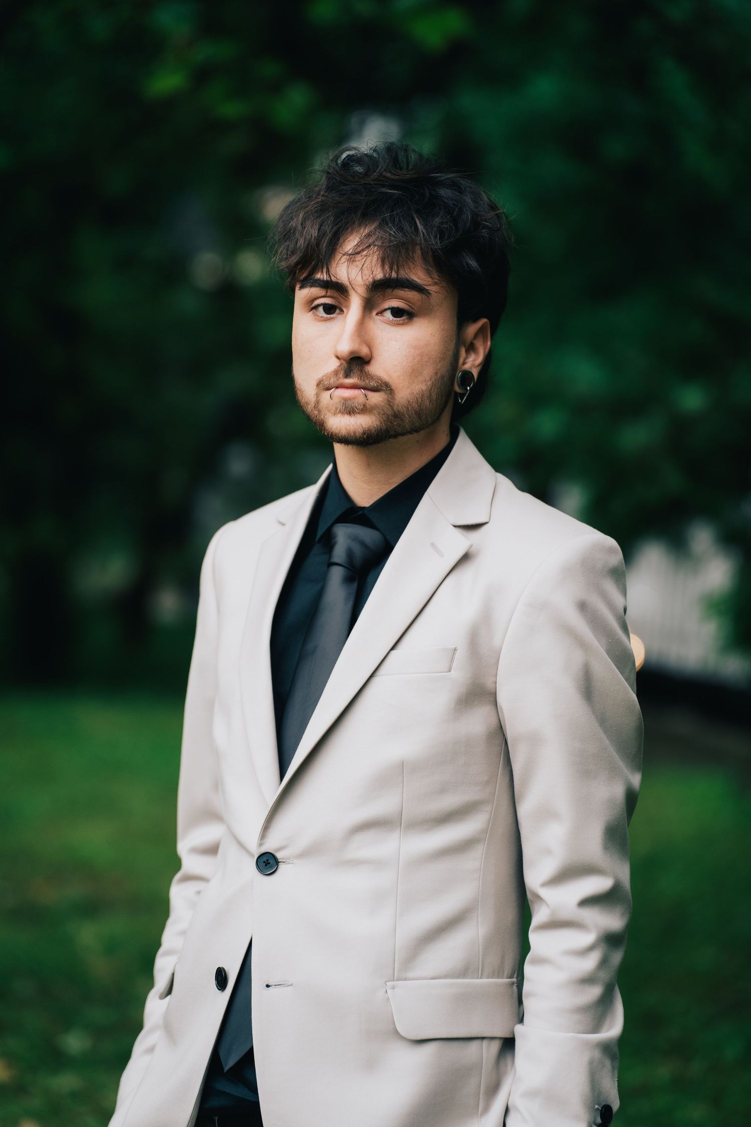 A young man with dark wavy hair and an earring poses in a beige blazer and dark tie on a grassy lawn surrounded by trees.