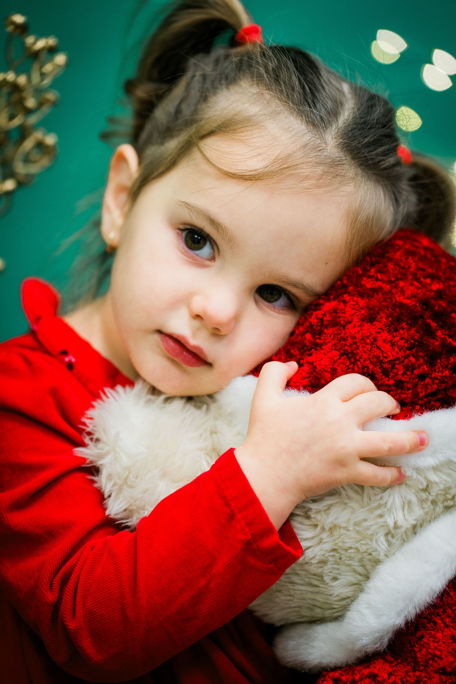 Toddler girl in a red top with pigtails rests her head on a plush red and white Santa hat against a green background.