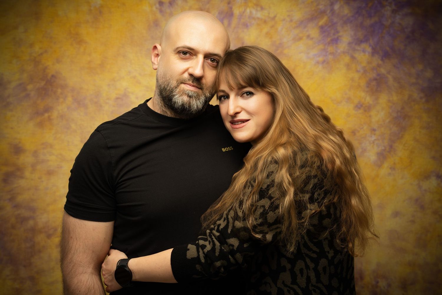 A bald bearded man in black embraces a smiling long-haired woman in a studio portrait with a golden painterly backdrop.