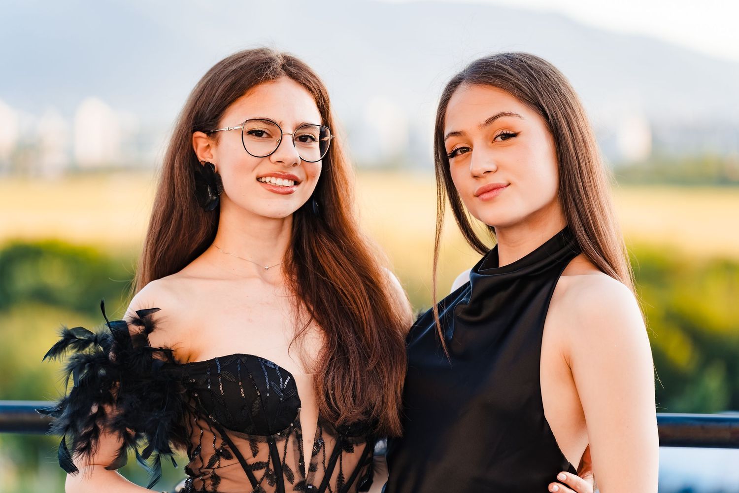 Two young women with long hair smile on a terrace, one in a black feathered lace top and one in a black halter dress.