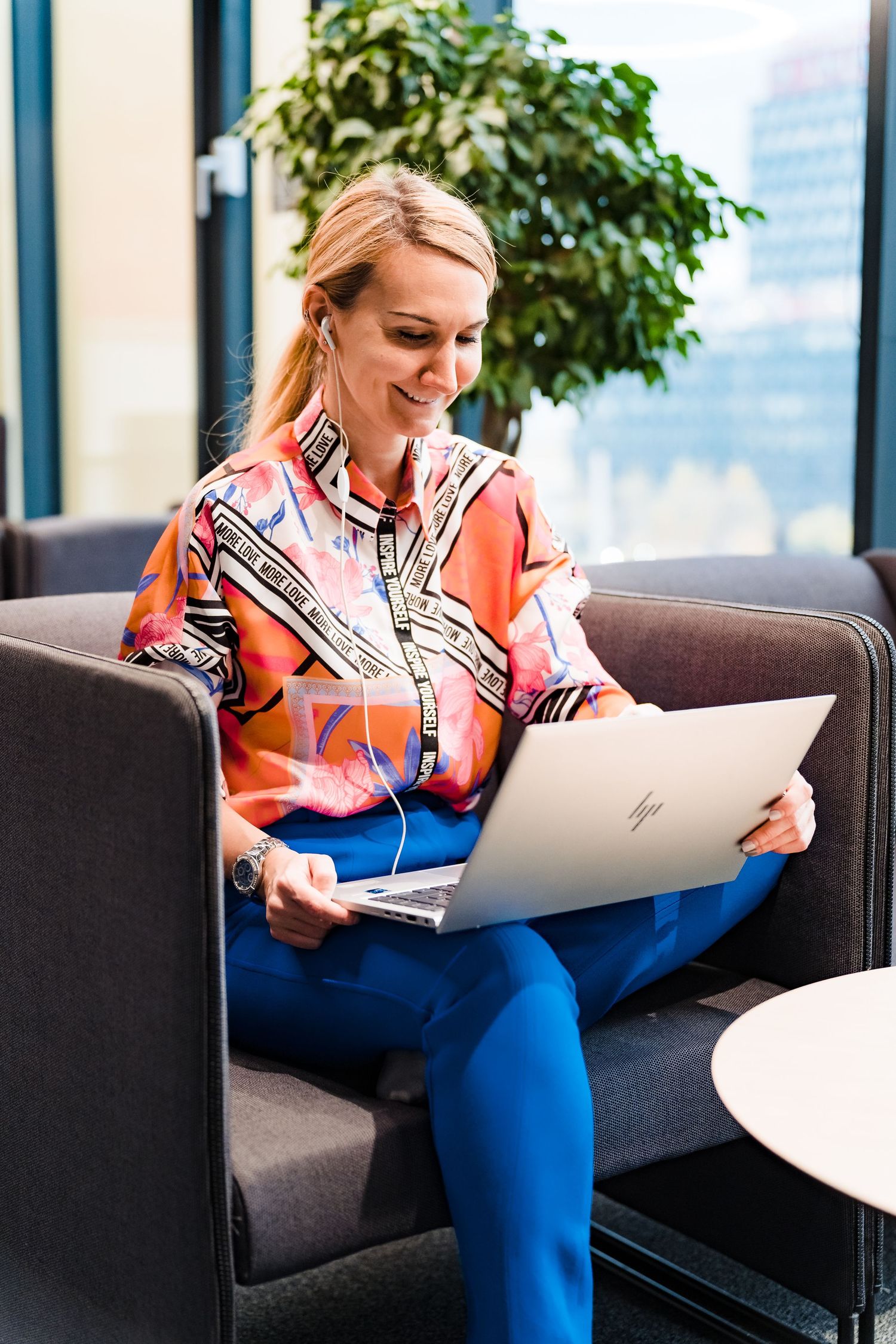Smiling woman in a vibrant orange blouse and blue pants works on a laptop while seated on a gray lounge sofa near a window.