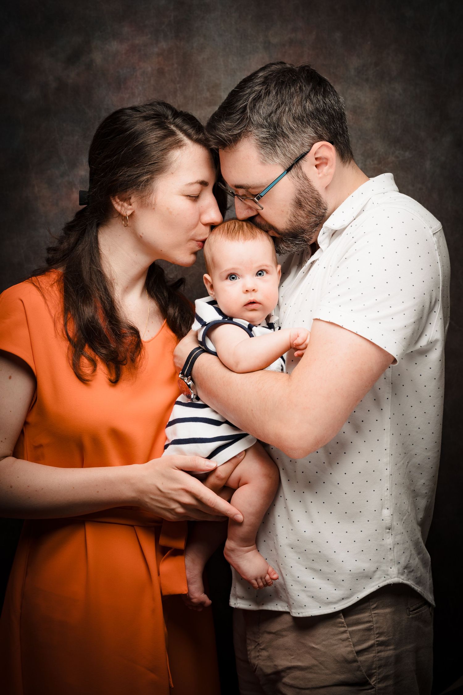 Woman in orange dress and bearded man in glasses kiss their baby's forehead in a warm family portrait against a dark textured backdrop.