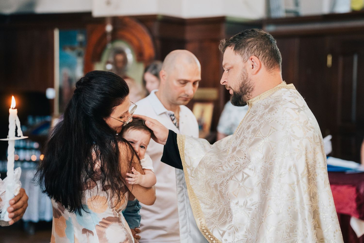 A priest in golden vestments performs a baptism blessing as a mother holds her baby and a godfather looks on beside a lit candle.
