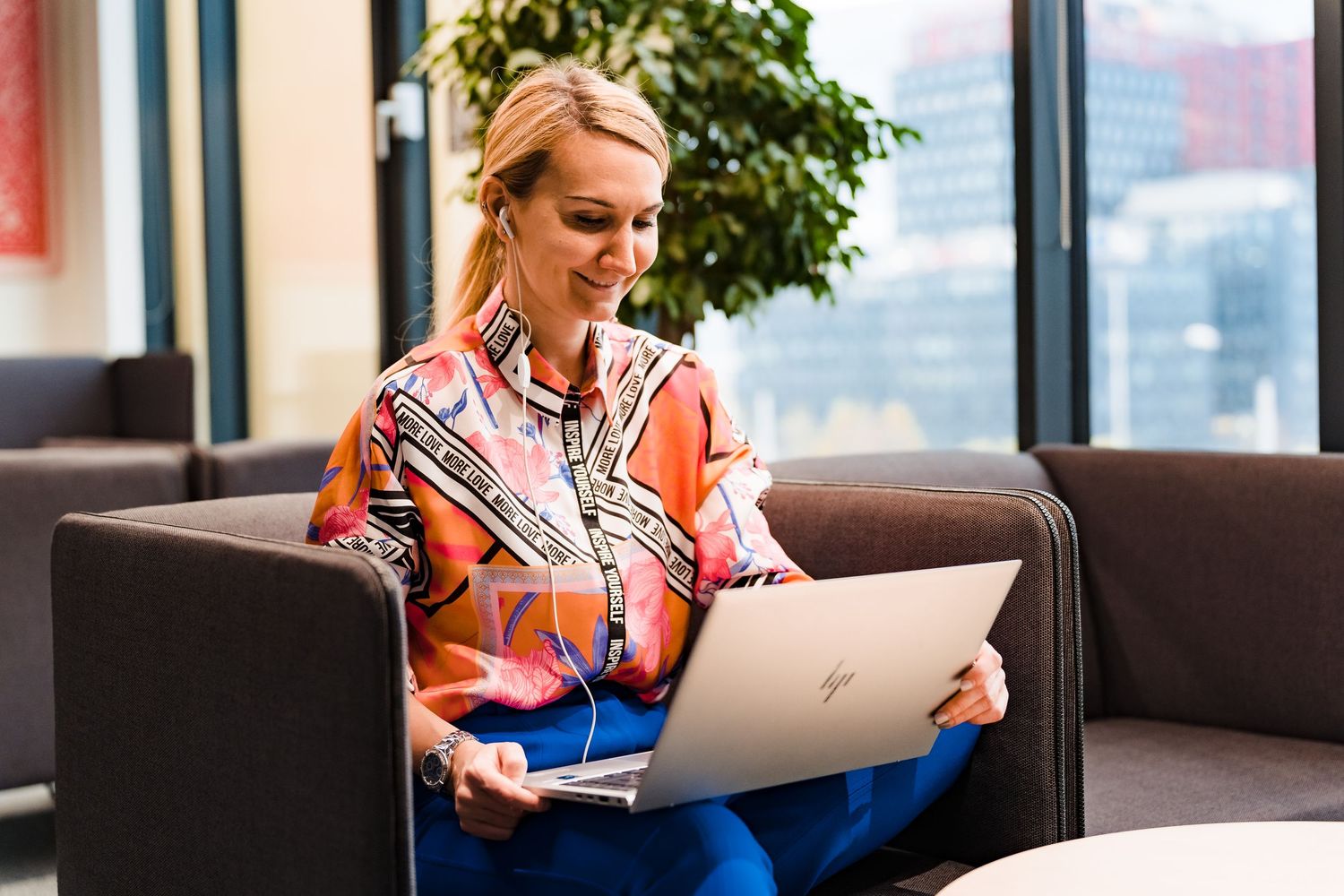 Happy blonde woman in a bold patterned blouse uses a laptop on a modern office lounge chair with city windows behind her.