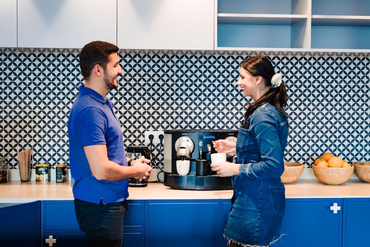 A man and a woman chat and laugh beside an espresso machine in a blue office kitchen with a patterned tile backsplash.