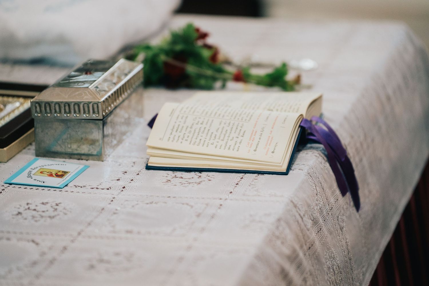 An open prayer book with a purple ribbon rests on a lace-covered table beside a silver box and red flowers at a baptism.