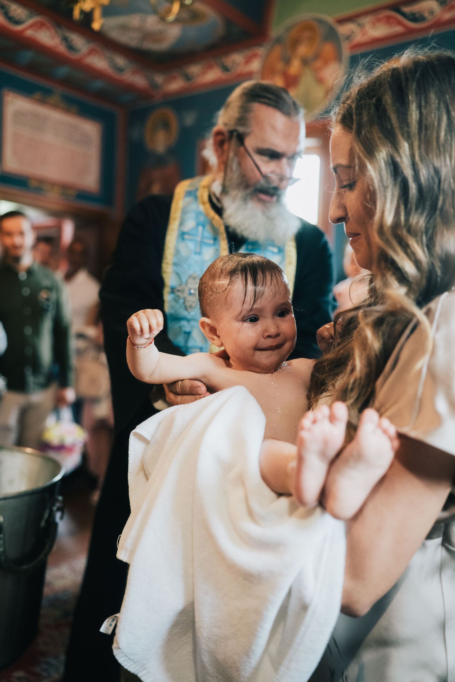 A bearded priest in blue vestments holds a smiling baby wrapped in a white towel during a baptism in an ornate church.