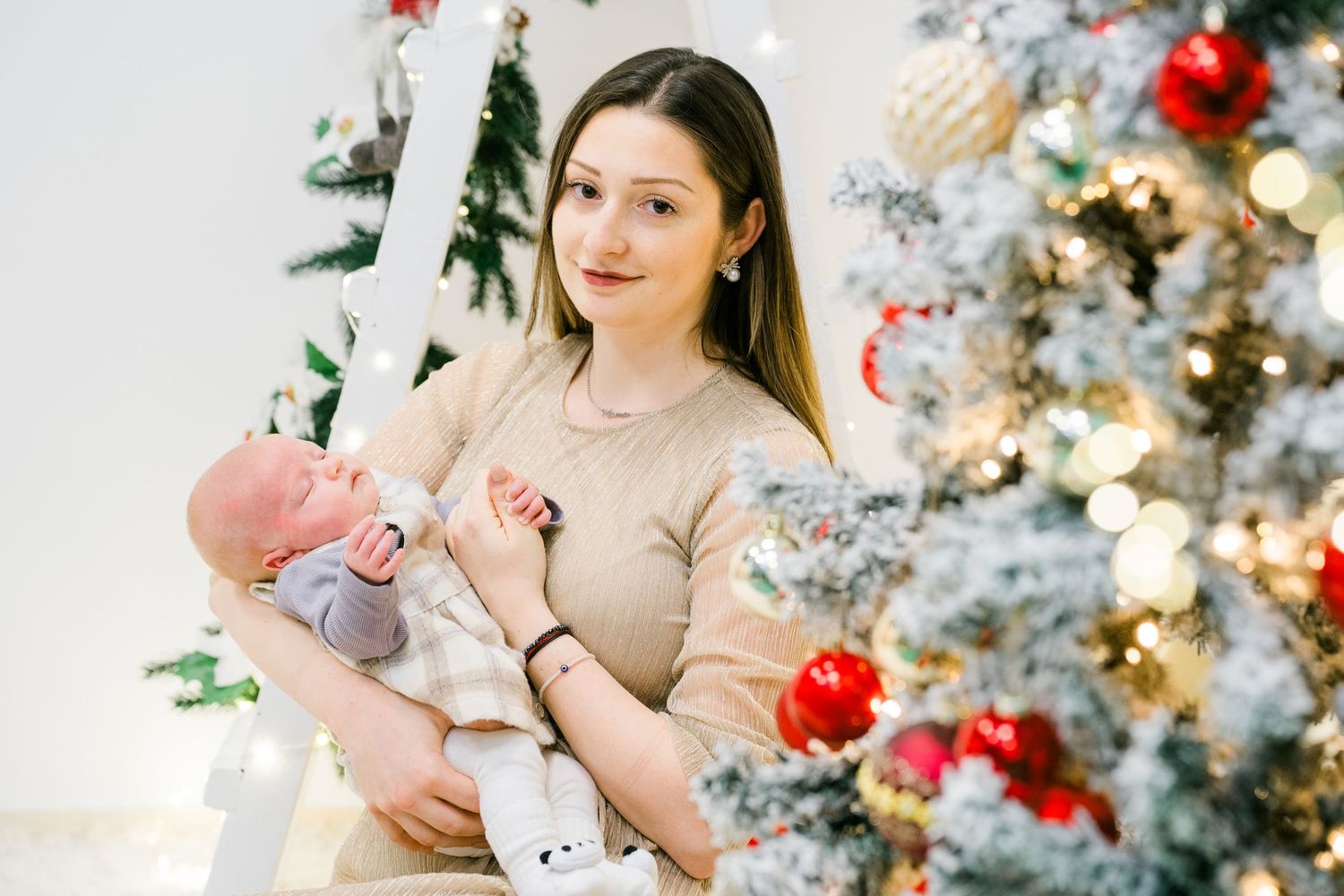 Smiling mother holds her newborn baby next to a snowy Christmas tree decorated with red and gold ornaments.