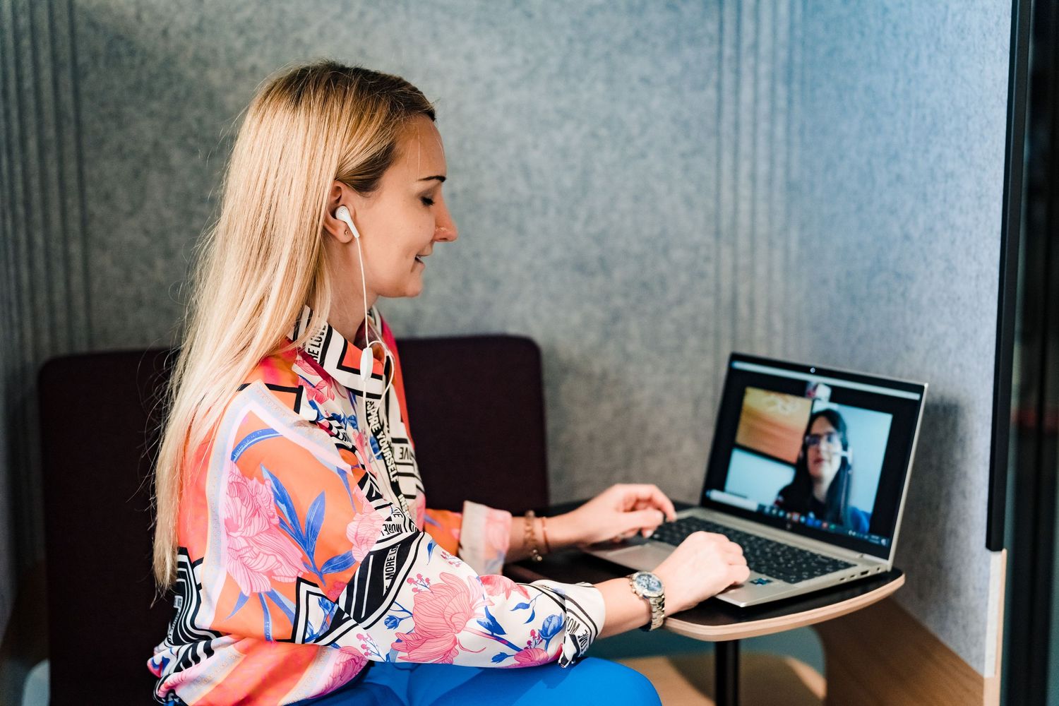 Blonde woman in a colorful patterned blouse wears earbuds and video chats on her laptop in a quiet, acoustic office nook.