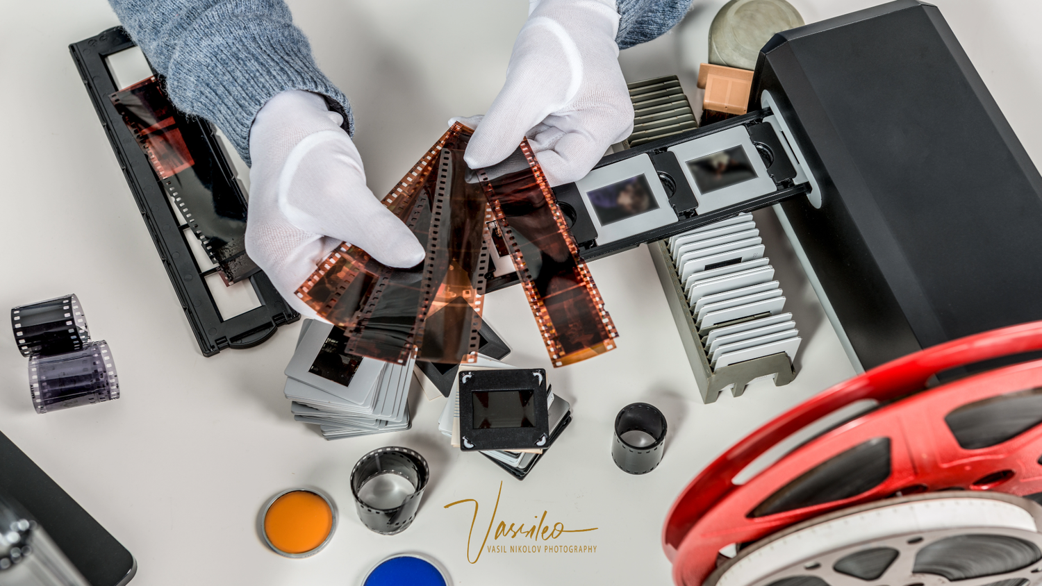 Gloved hands examine strips of developed film negatives surrounded by slide mounts, film reels, and scanning equipment on a white surface.