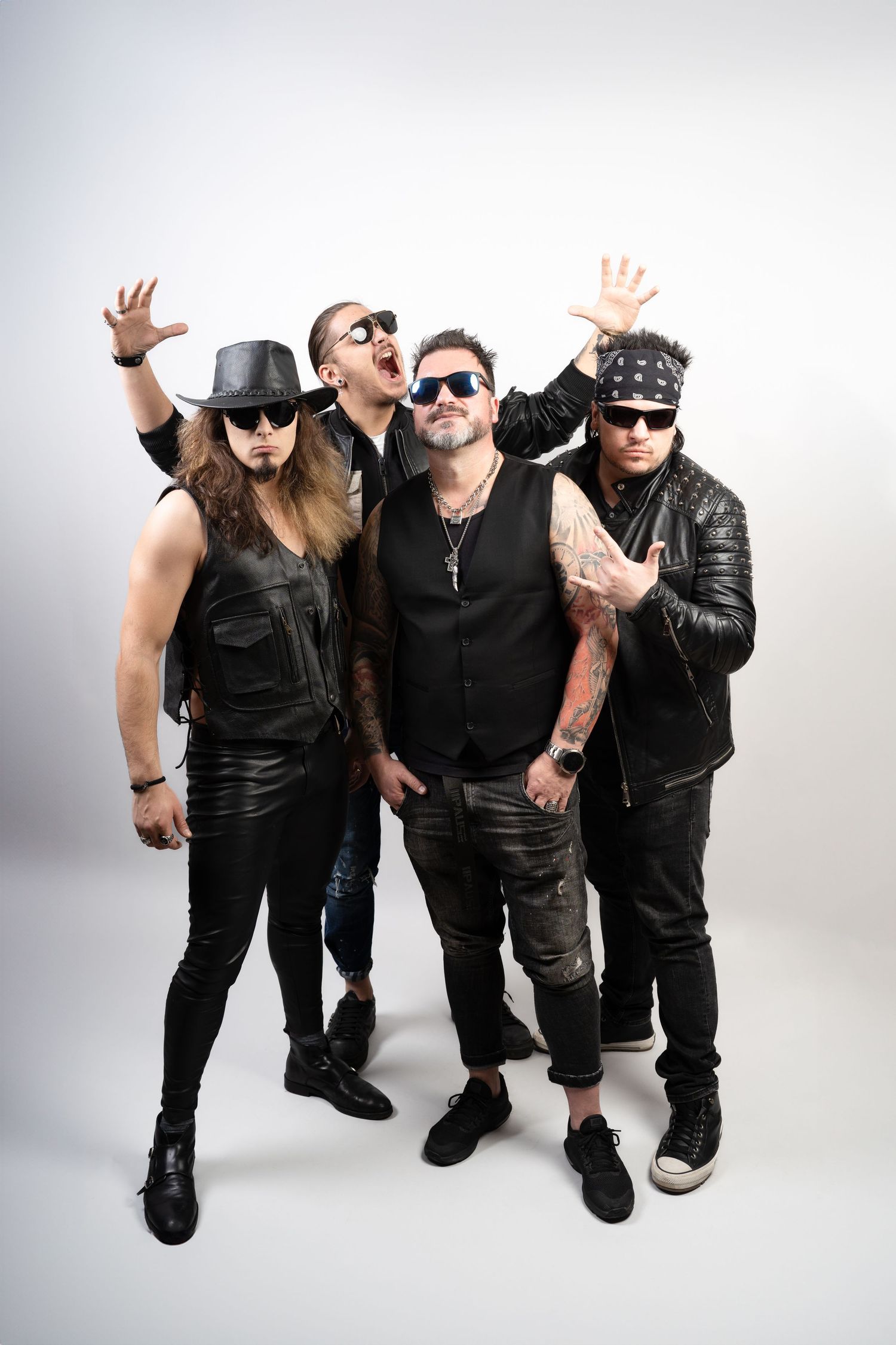 Four rock band members in black leather outfits, sunglasses, and tattoos pose with energetic gestures against a plain studio backdrop.