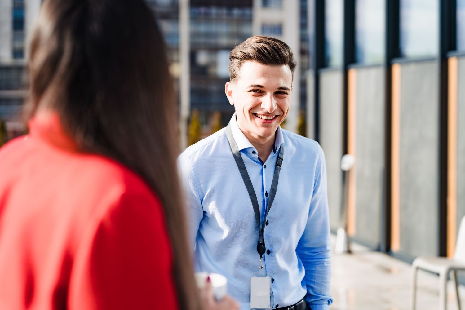 Smiling man in a light blue shirt with a lanyard chats with a woman in red on an outdoor office terrace.