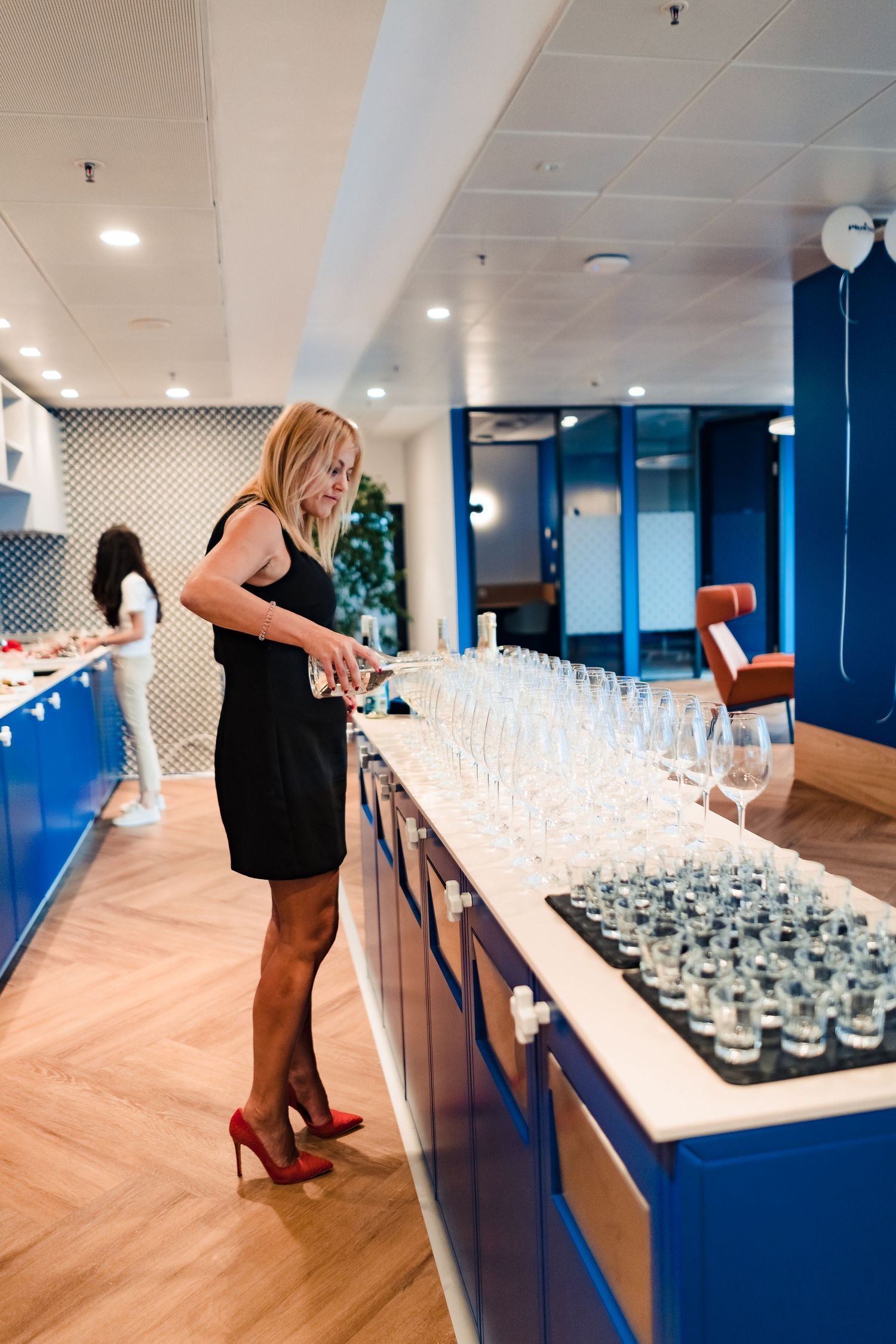 Woman in a black dress and red heels pours drinks at a long bar counter lined with glasses in a modern blue and white event space.
