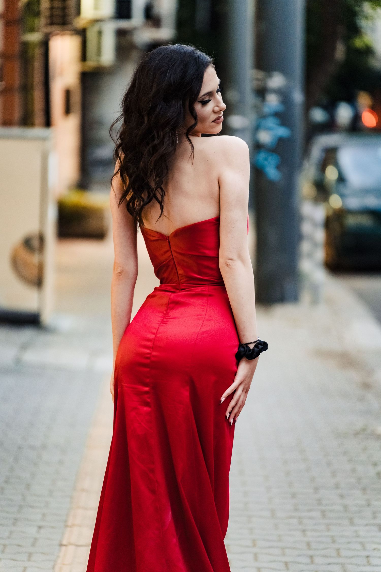 Woman in a long red satin gown walks down a paved city sidewalk past graffiti and parked cars.