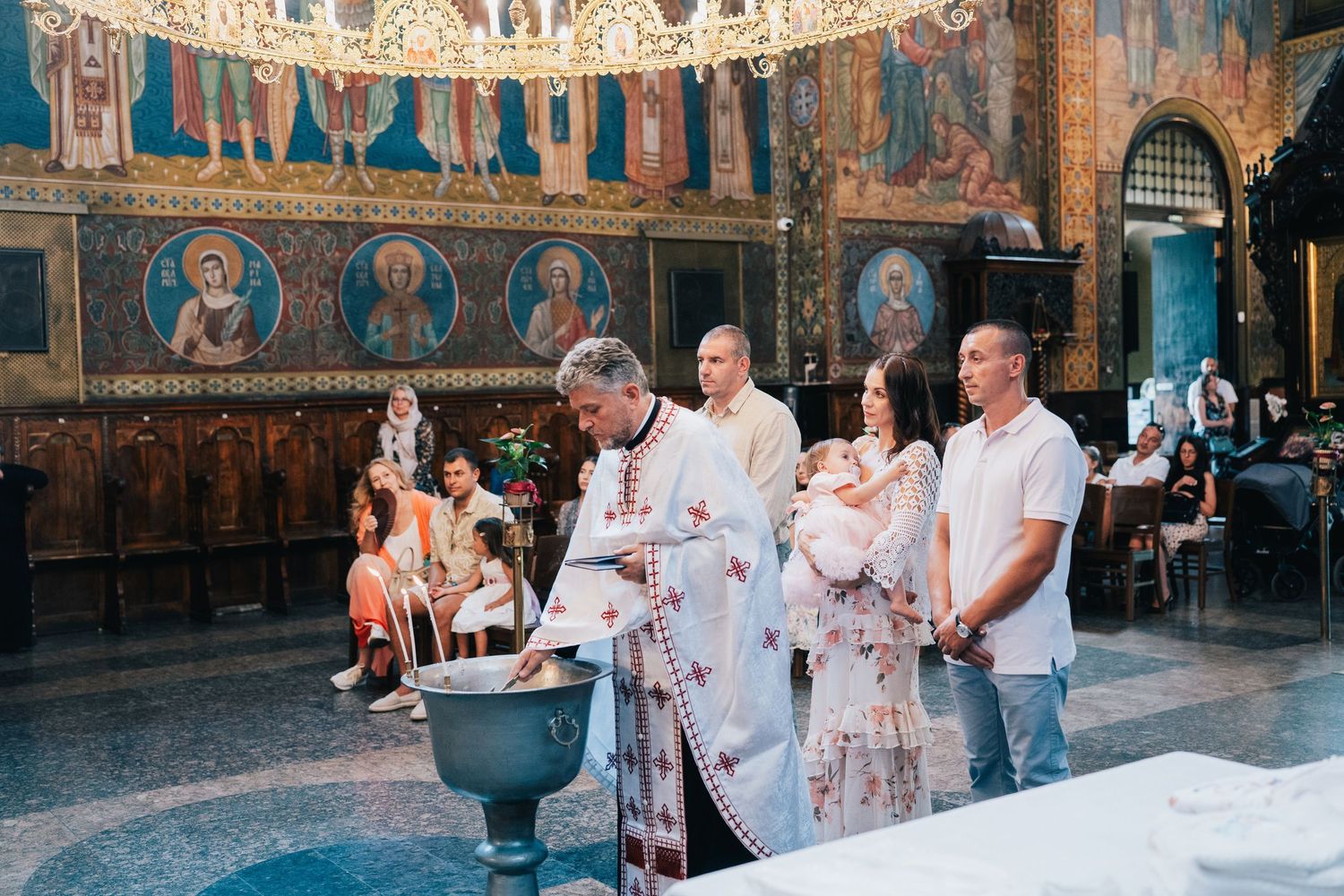 Priest in white vestments leads a christening at the baptismal font as parents hold their baby in a church decorated with icons.
