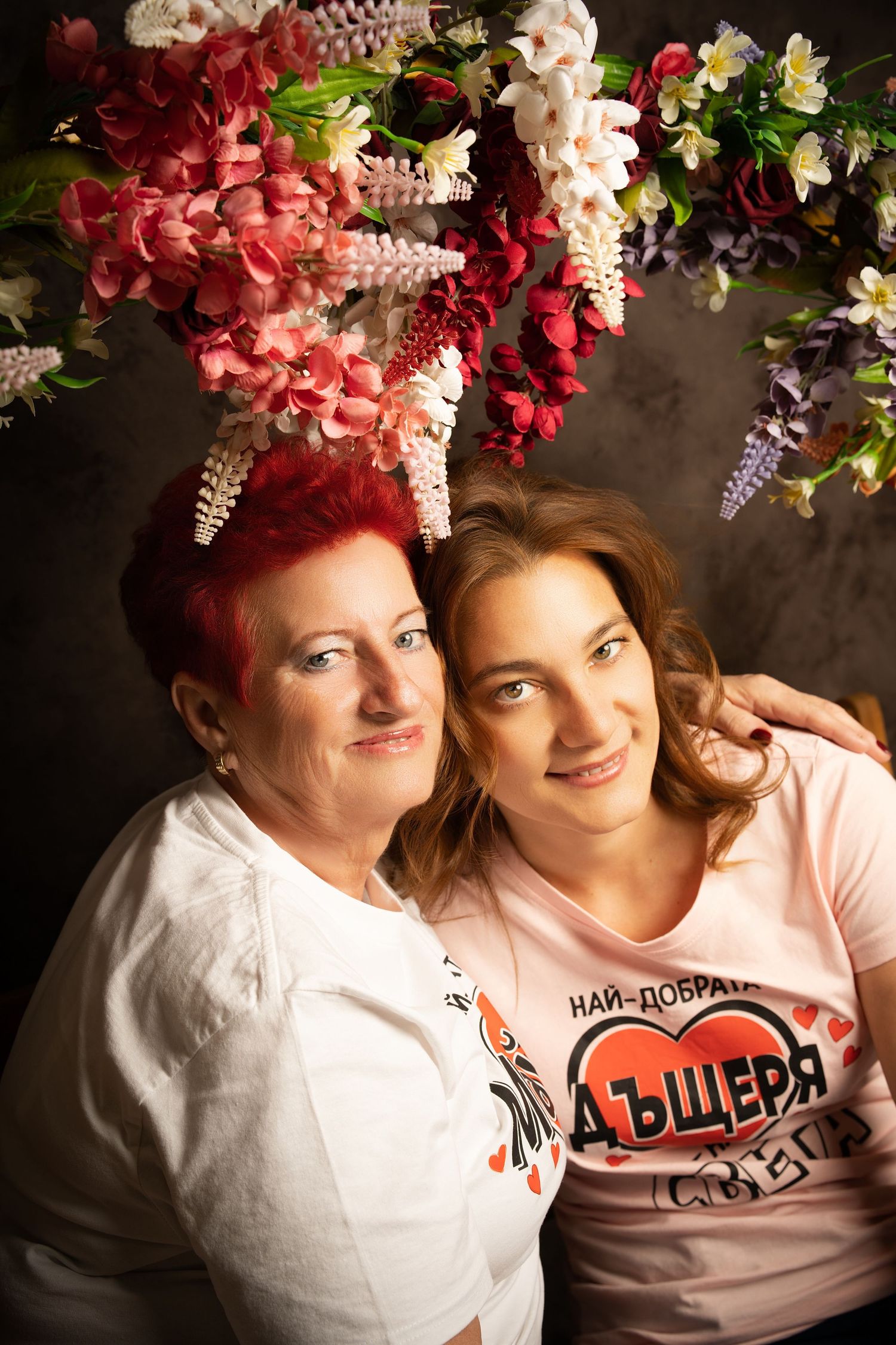 A mother and daughter in matching shirts hug closely and smile in a studio portrait framed by colorful hanging flowers.