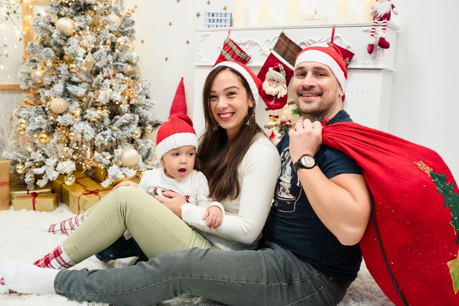 Happy family in Santa hats sits with their baby near a decorated Christmas tree, stockings and a red gift sack.