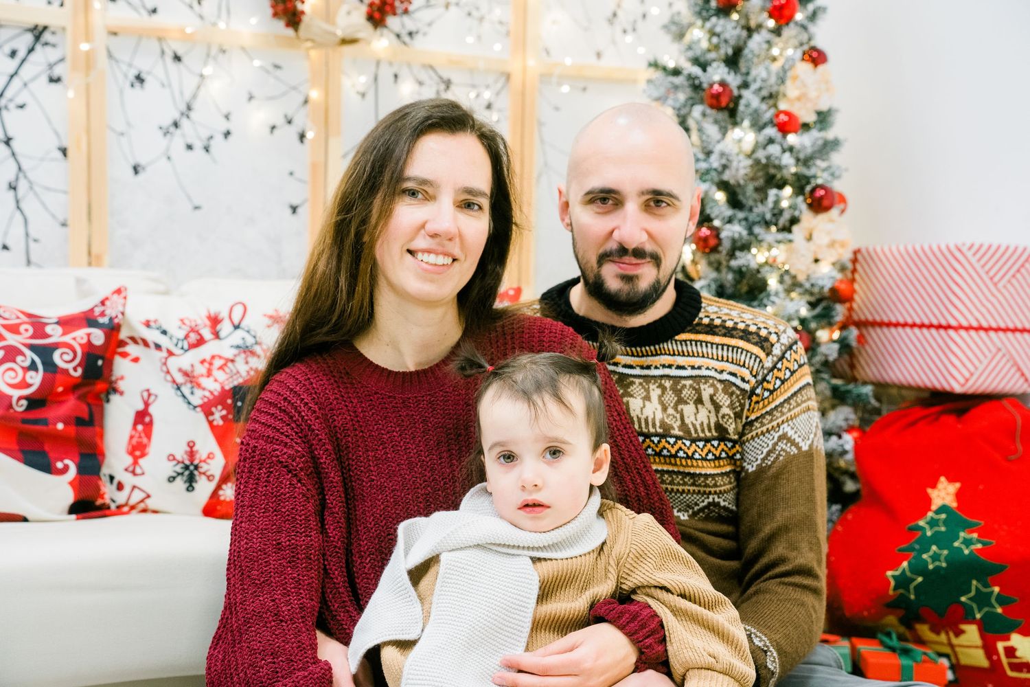 Mother, father and toddler smile together in a festive Christmas photo with a decorated tree and gift boxes behind them.