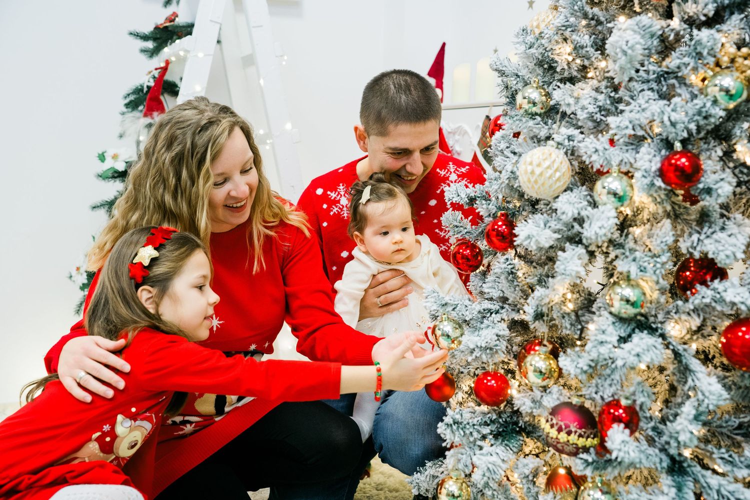 Family with two young daughters and a baby decorates a frosted Christmas tree with ornaments in matching red holiday outfits.