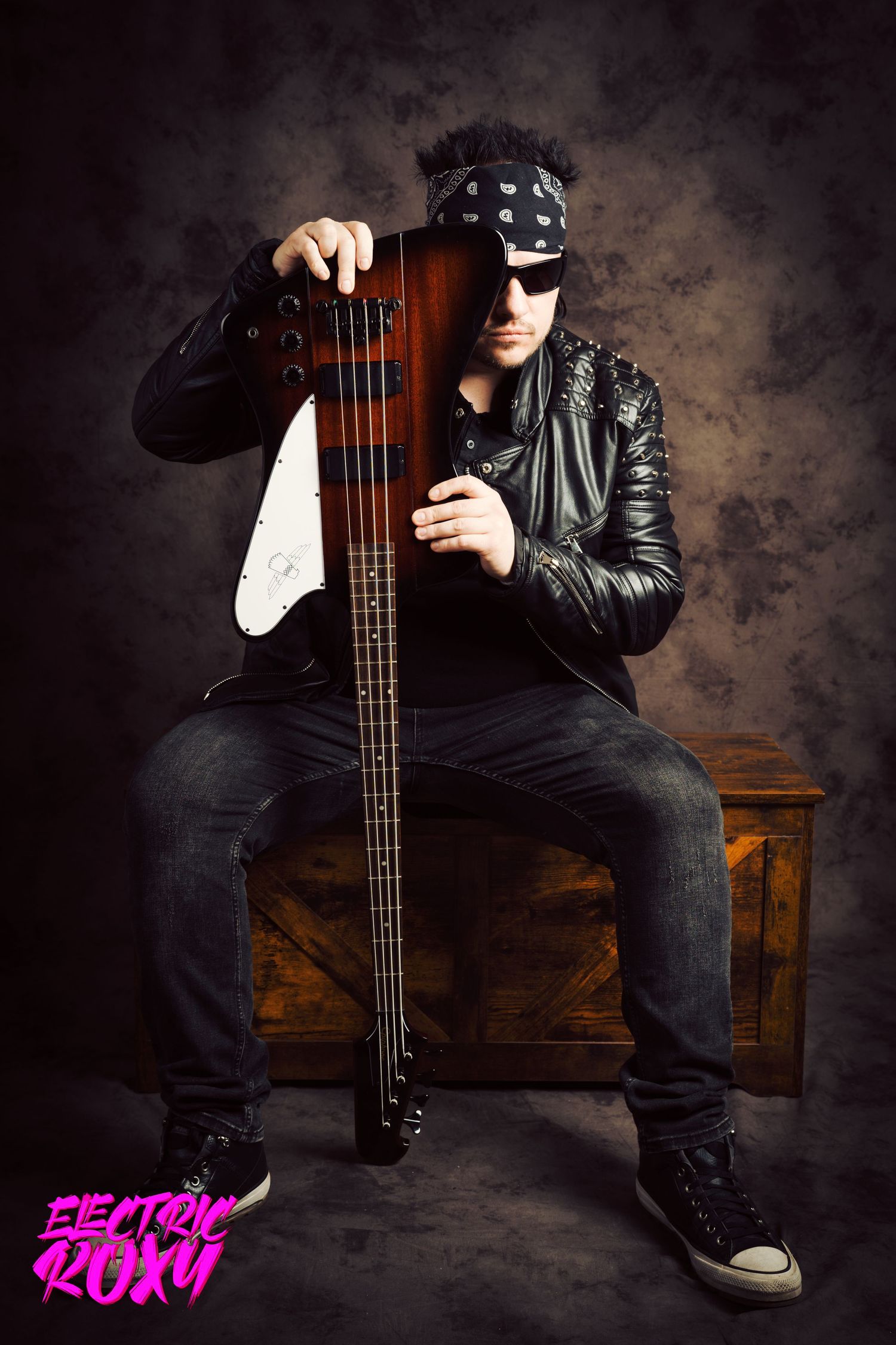 Bassist in sunglasses, bandana and studded leather jacket sits on a wooden crate holding a sunburst bass guitar in a moody studio portrait.