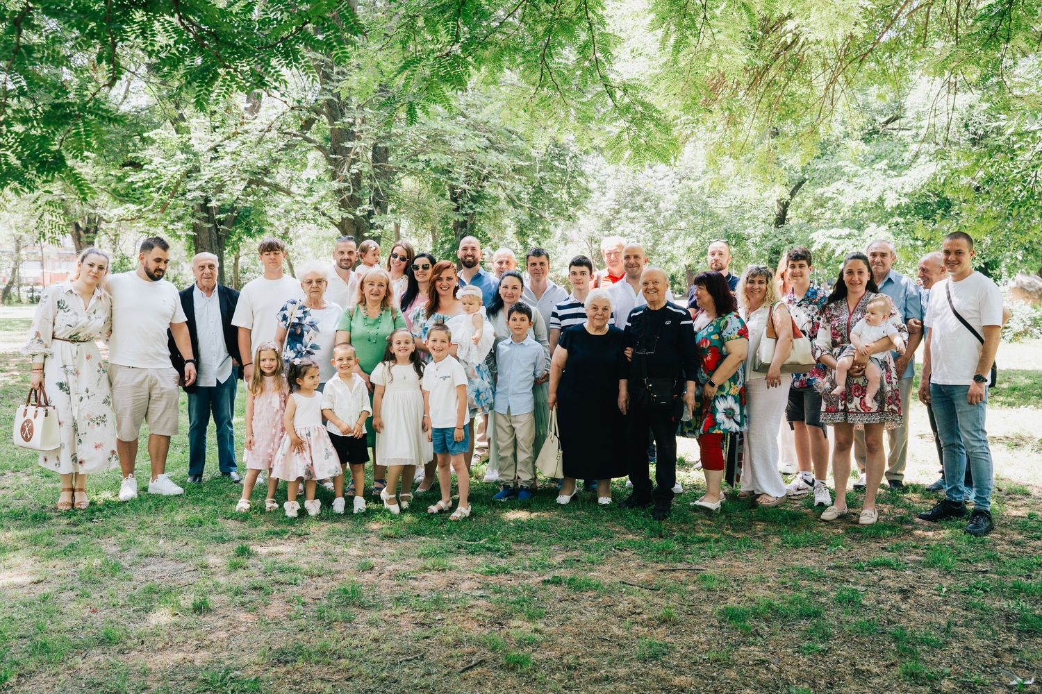 A large extended family and friends pose together for a group photo on a grassy lawn under green trees after a baptism celebration.
