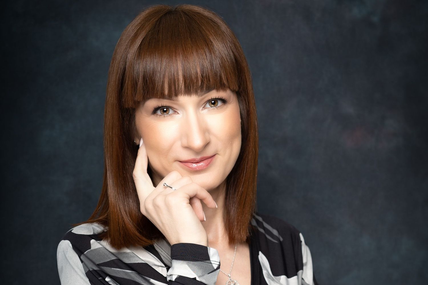 Woman with long auburn hair and bangs poses with her hand at her cheek, wearing a black and white striped top against a dark backdrop.